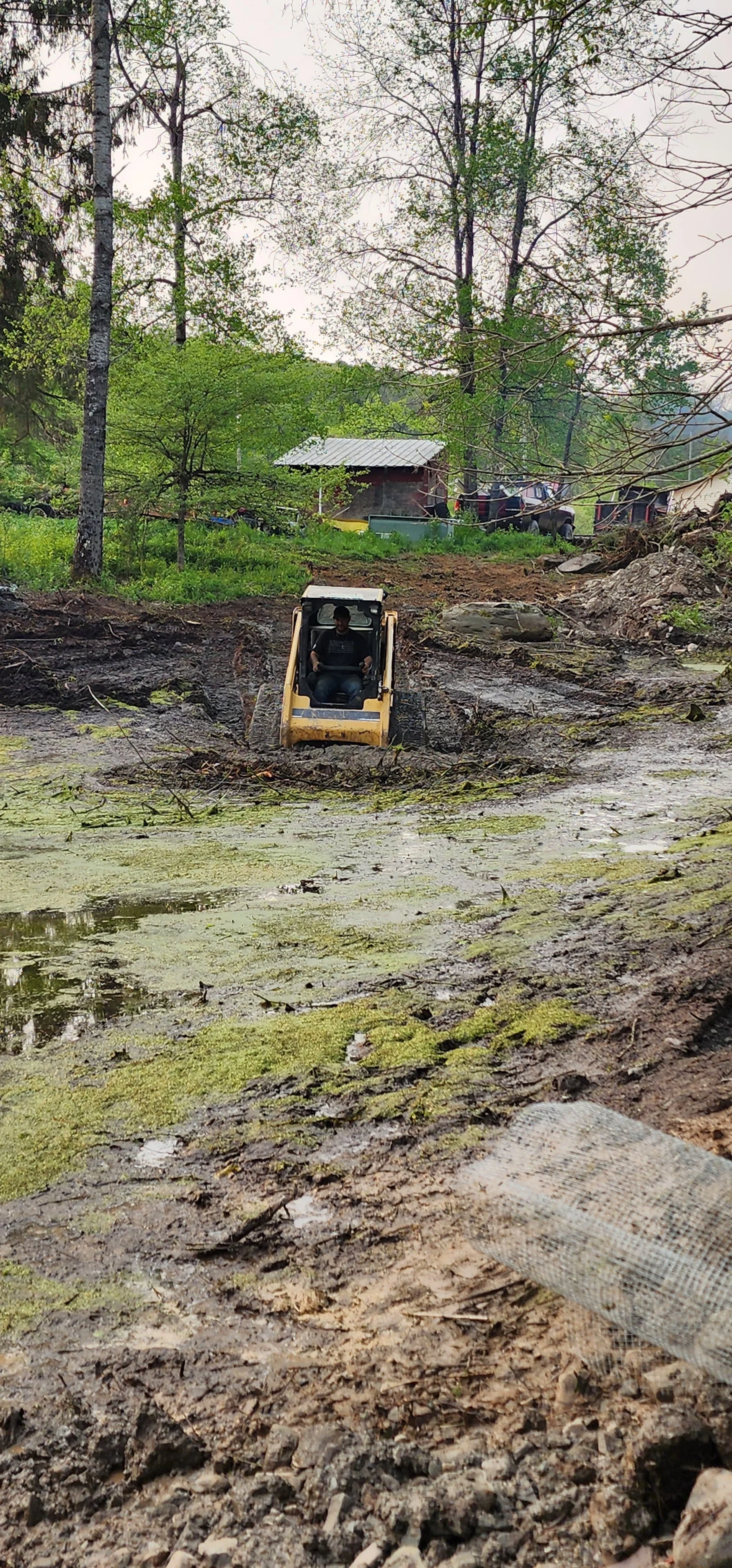 A person operating a small excavator in a muddy, waterlogged area with green moss and swampy water, surrounded by trees and a rural landscape in the background.