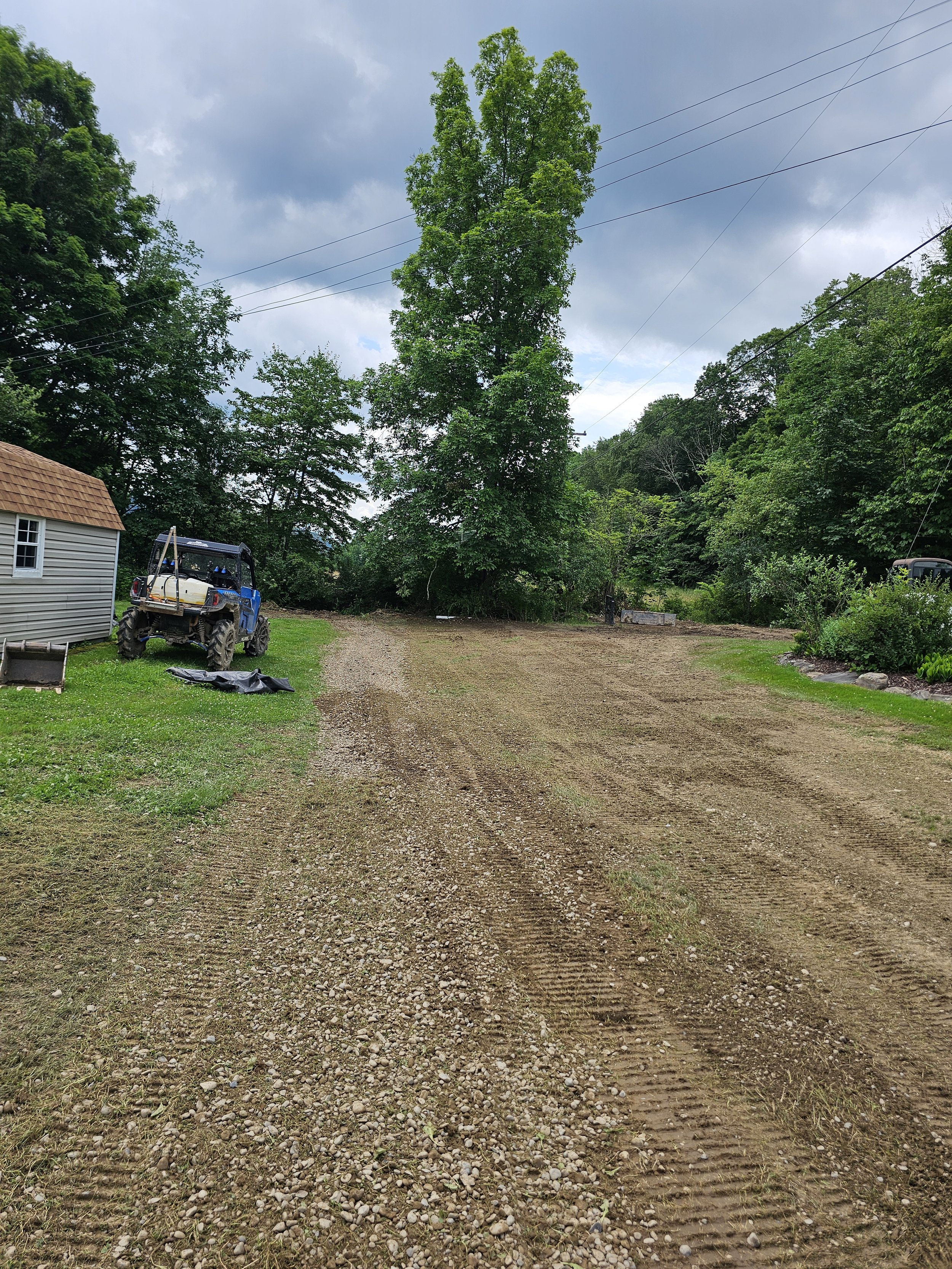 A rural backyard with a gravel driveway, a small shed, a blue all-terrain vehicle, a large leafy tree, and cloudy sky.