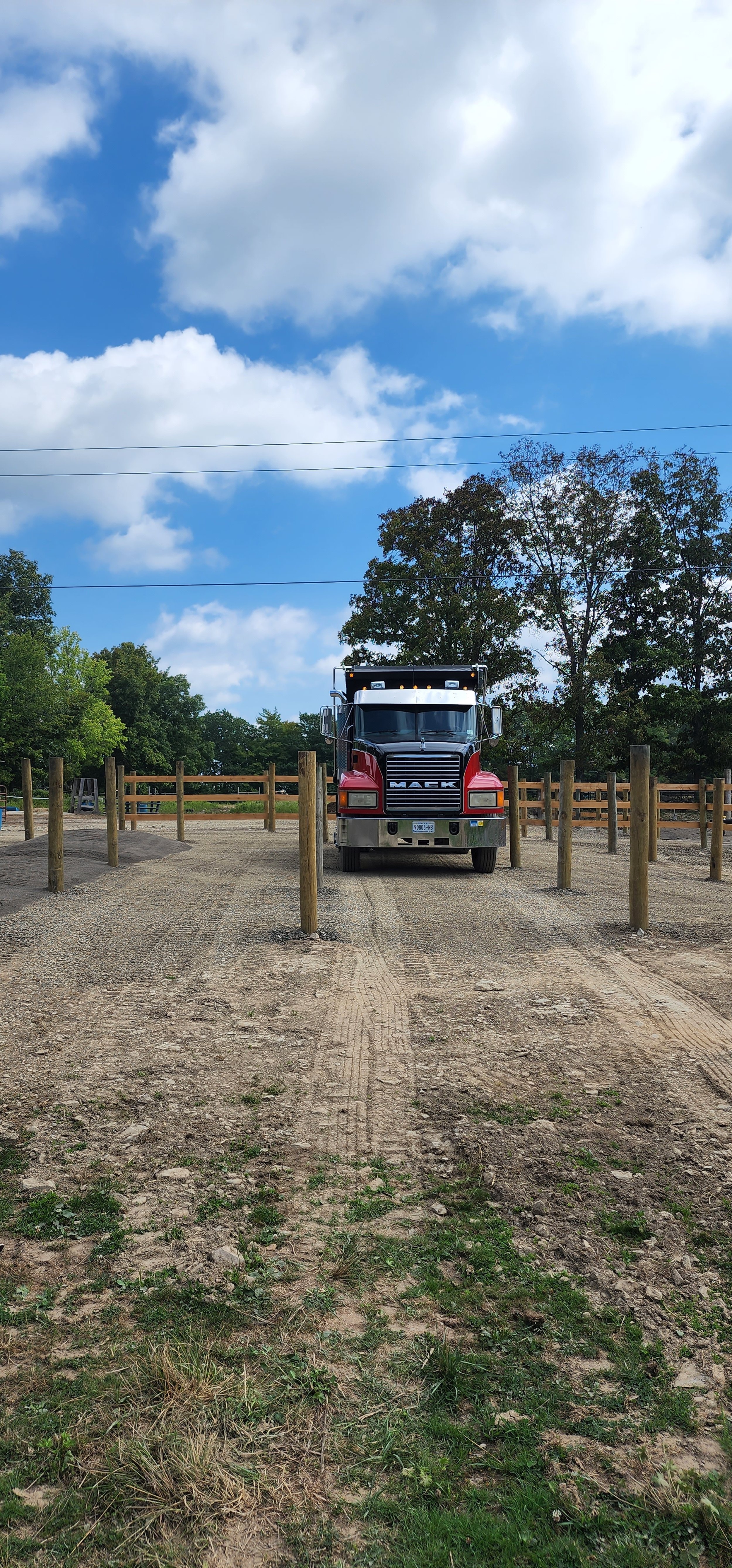 A red Mack truck parked in a gravel lot with wooden posts. There are trees and a blue sky with clouds in the background.