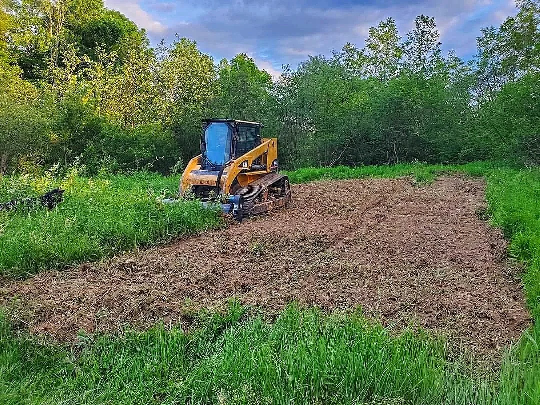 A small bulldozer working on clearing land, surrounded by green trees and grass, under a partly cloudy sky.