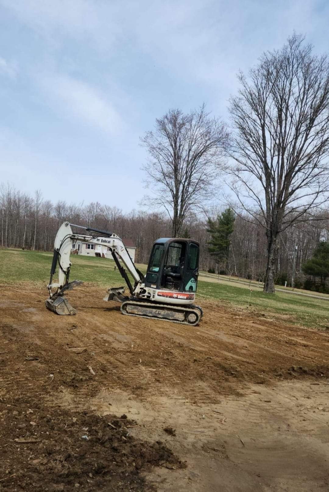 A small white and black excavator on a dirt lot, with a grassy field and leafless trees in the background under a cloudy sky.