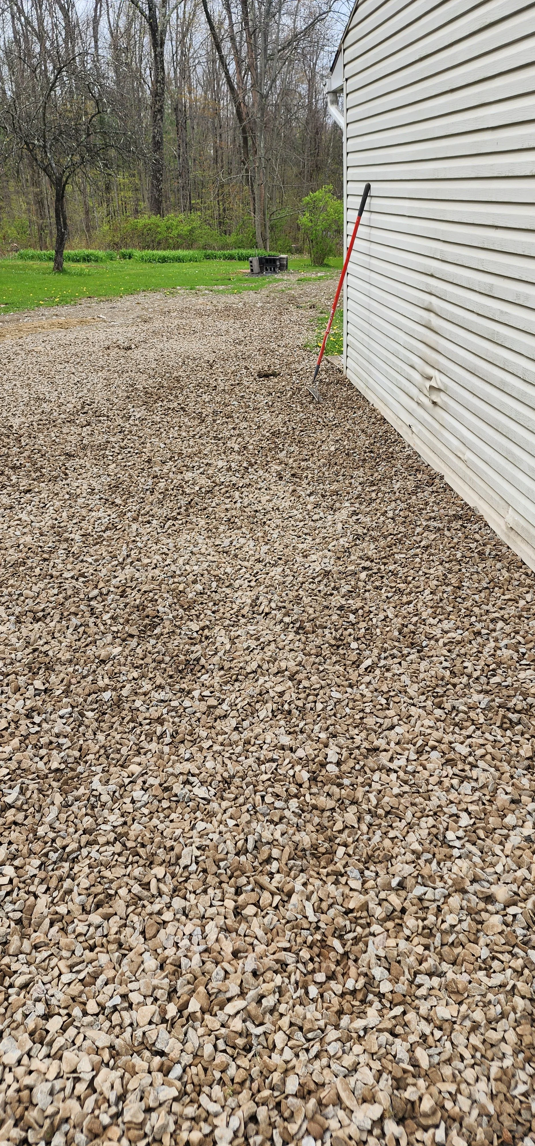 Gravel driveway next to a white house with a rake leaning against the wall, and a backyard with grass and trees in the background.