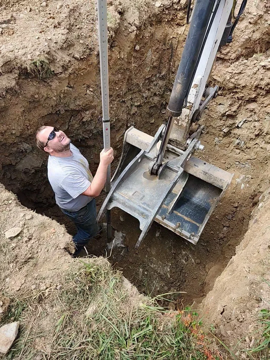 A man in a gray t-shirt, sunglasses, and dark pants is standing in a large excavation site with dirt walls, holding a measuring rod and operating an excavator arm.
