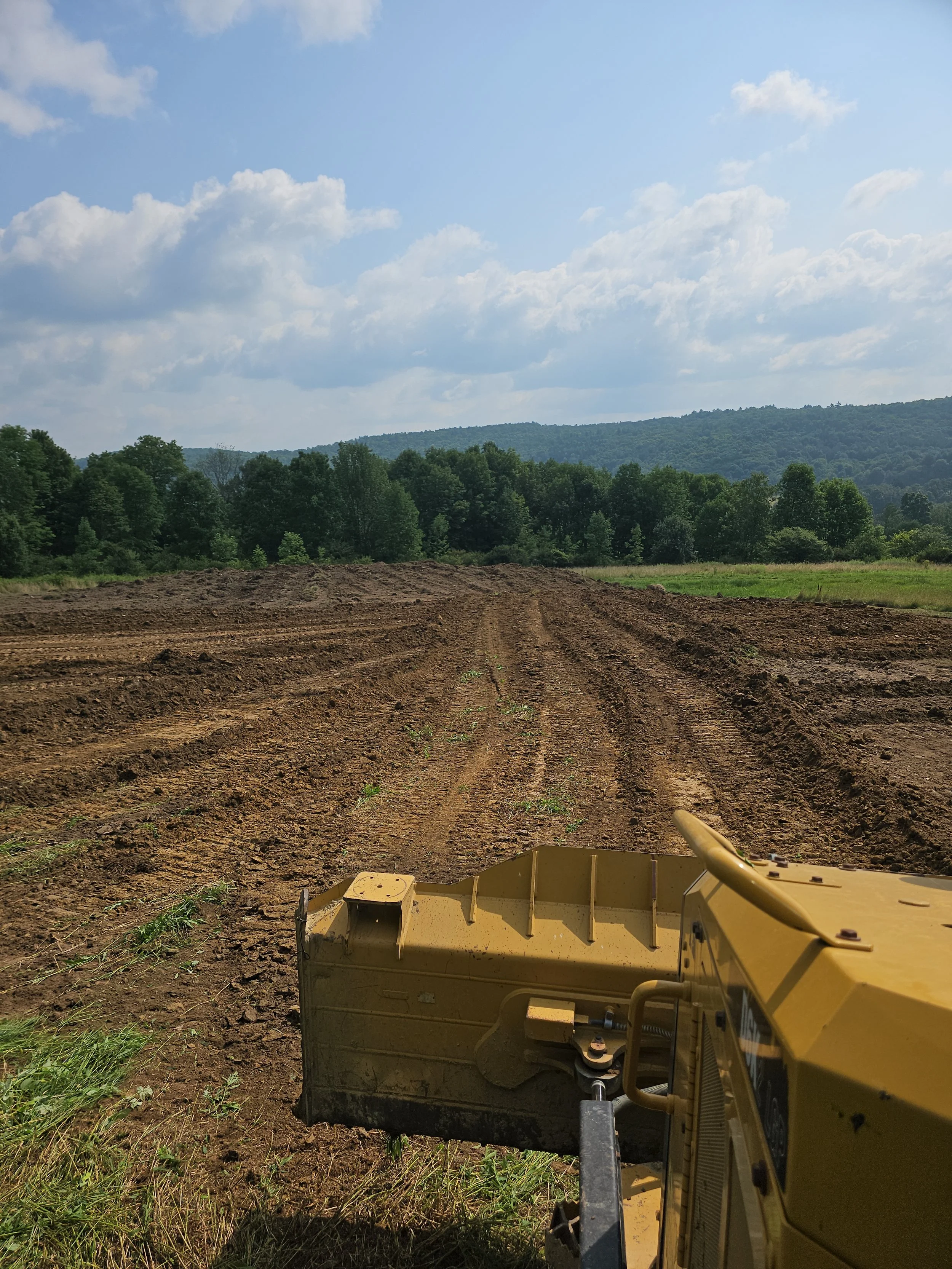 View of a freshly plowed field with a tractor blade in the foreground, surrounded by trees and distant hills under a partly cloudy sky.