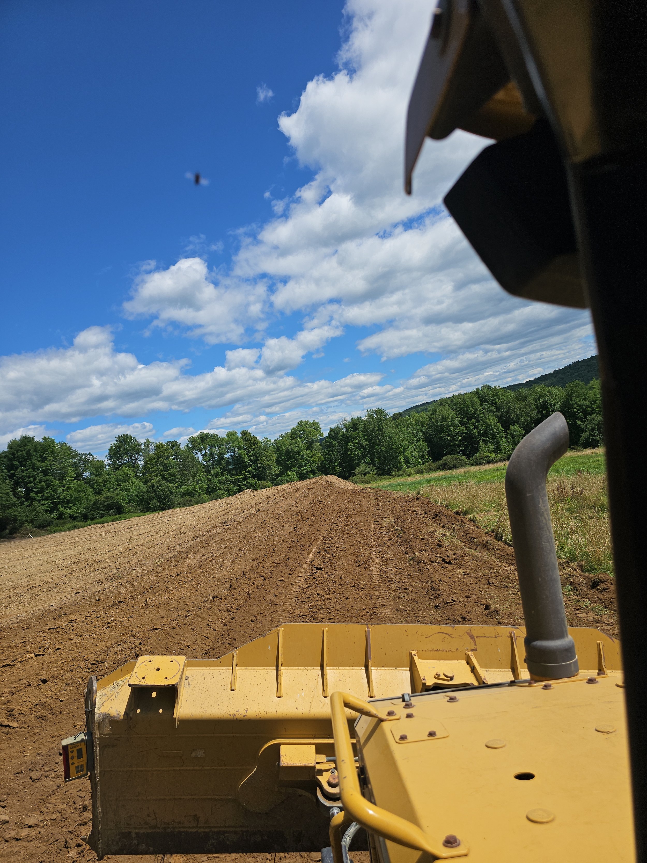 View from a yellow bulldozer on a dirt road with green trees and a blue sky with clouds in the background.