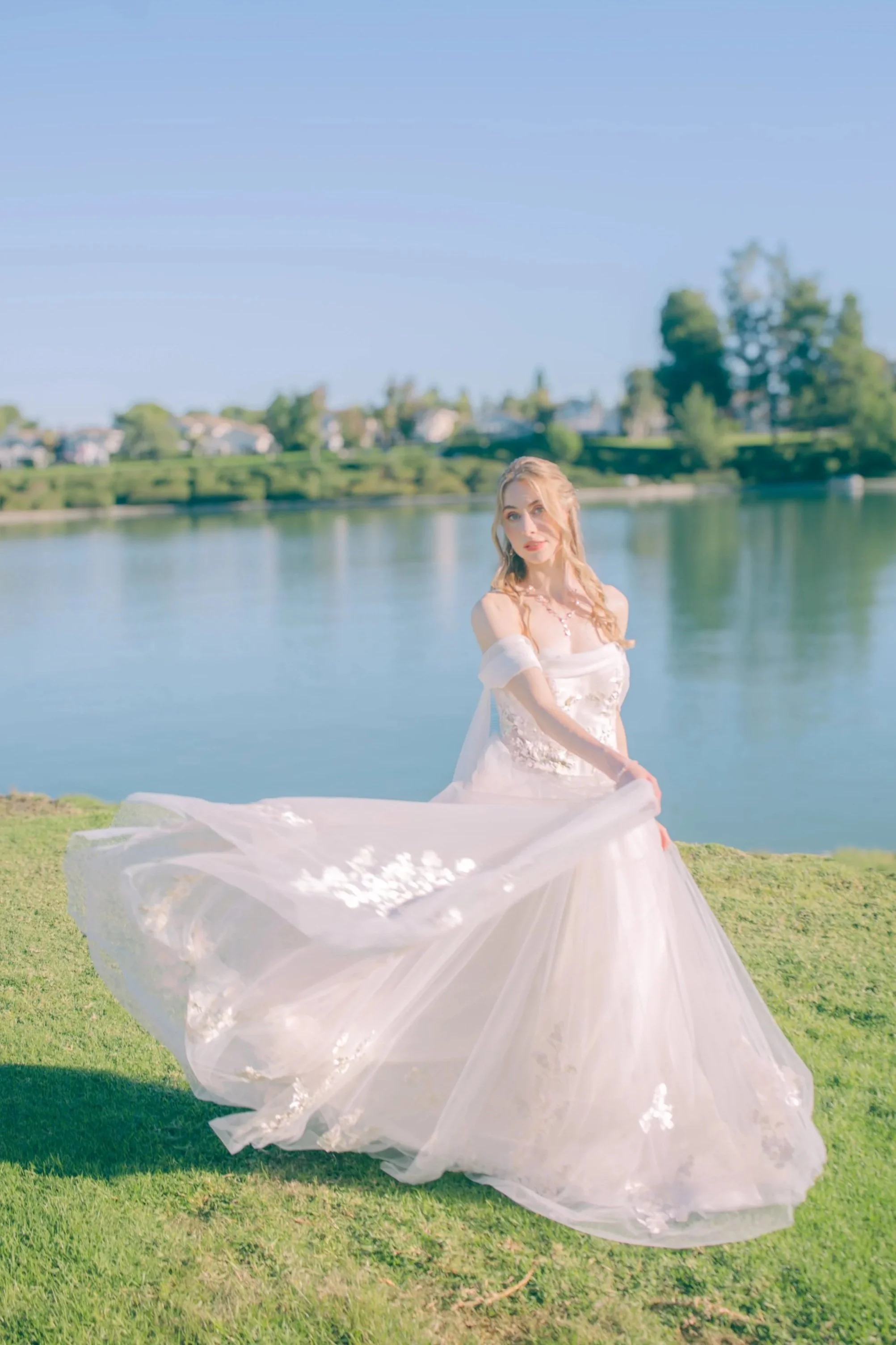 A young woman in a white and blush wedding dress standing on grass near a body of water on a sunny day.