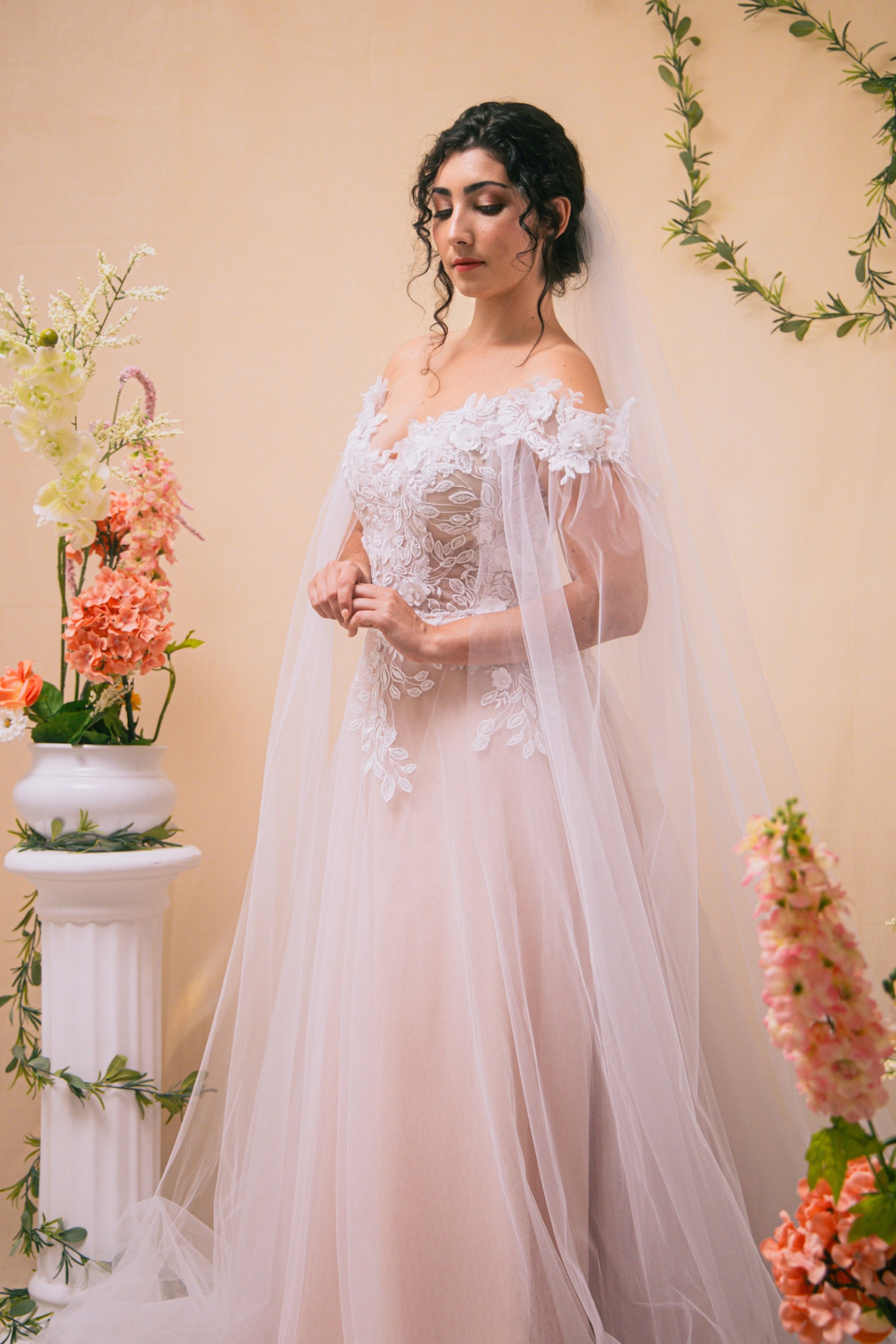 A woman in an off-the-shoulder white wedding dress with lace details, standing among floral arrangements and greenery, with her eyes closed and hands clasped.