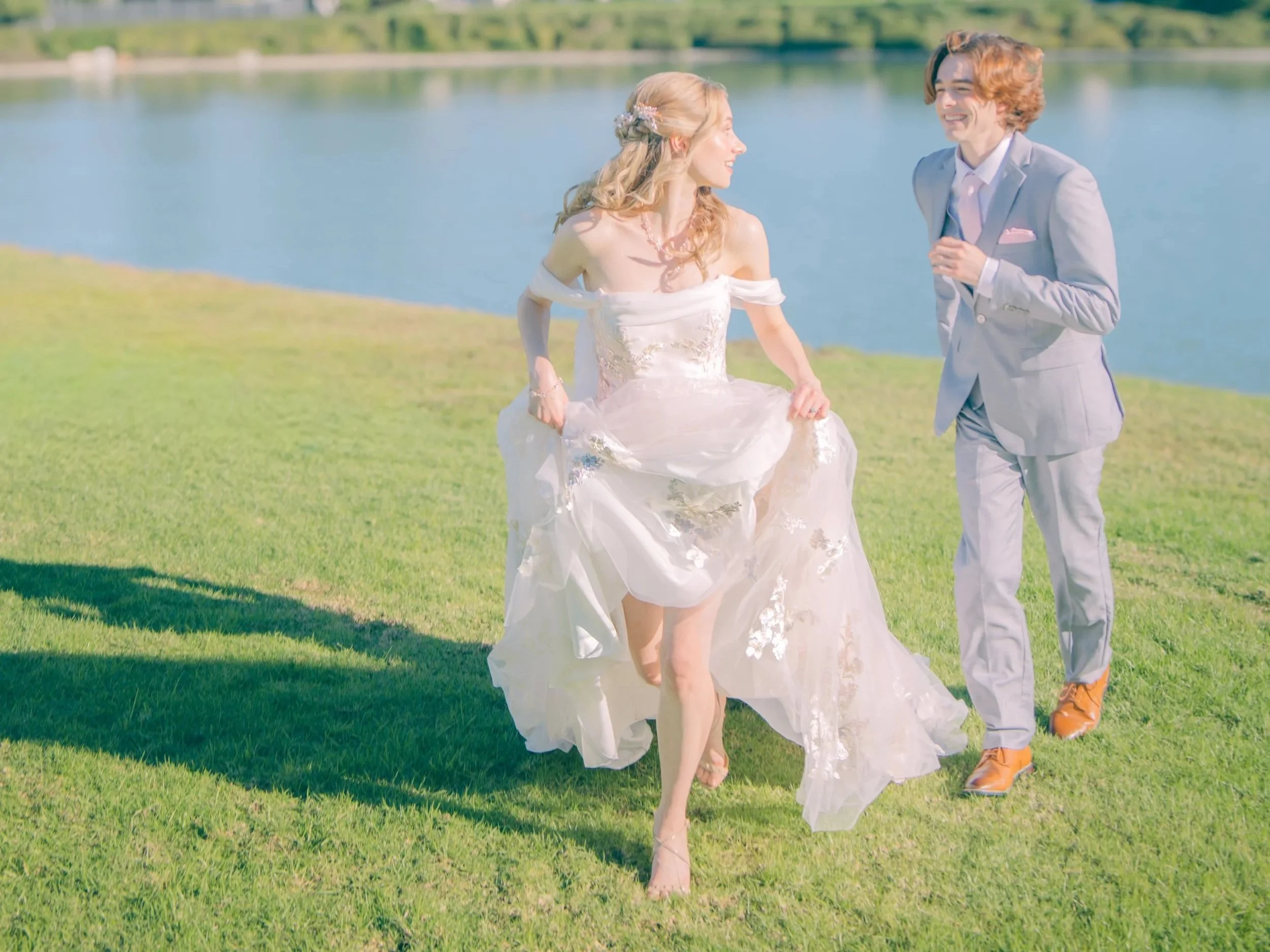 A woman in a wedding dress and a man in a gray suit are outdoors near a body of water, smiling and laughing together.