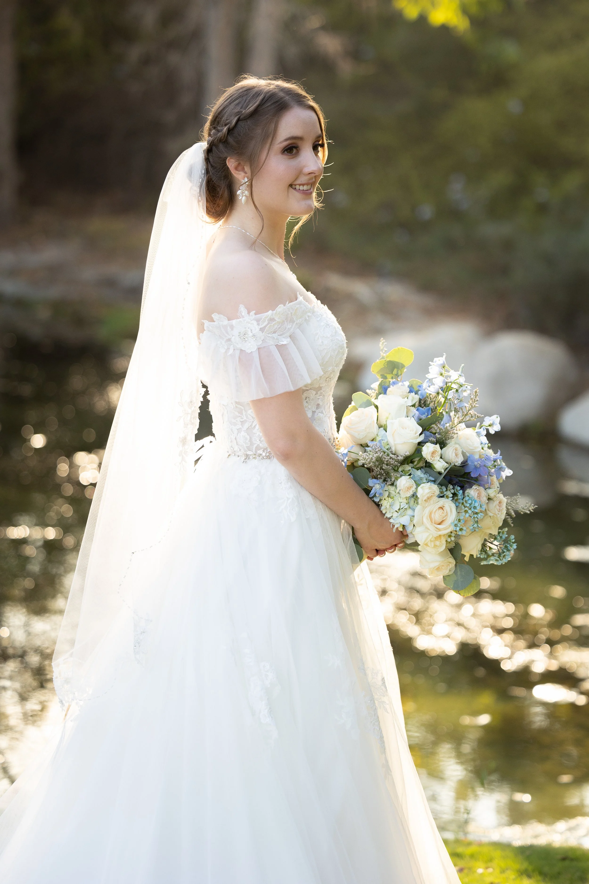 A smiling bride stands outdoors near a body of water, holding a bouquet of white and blue flowers, wearing a white wedding dress and veil.