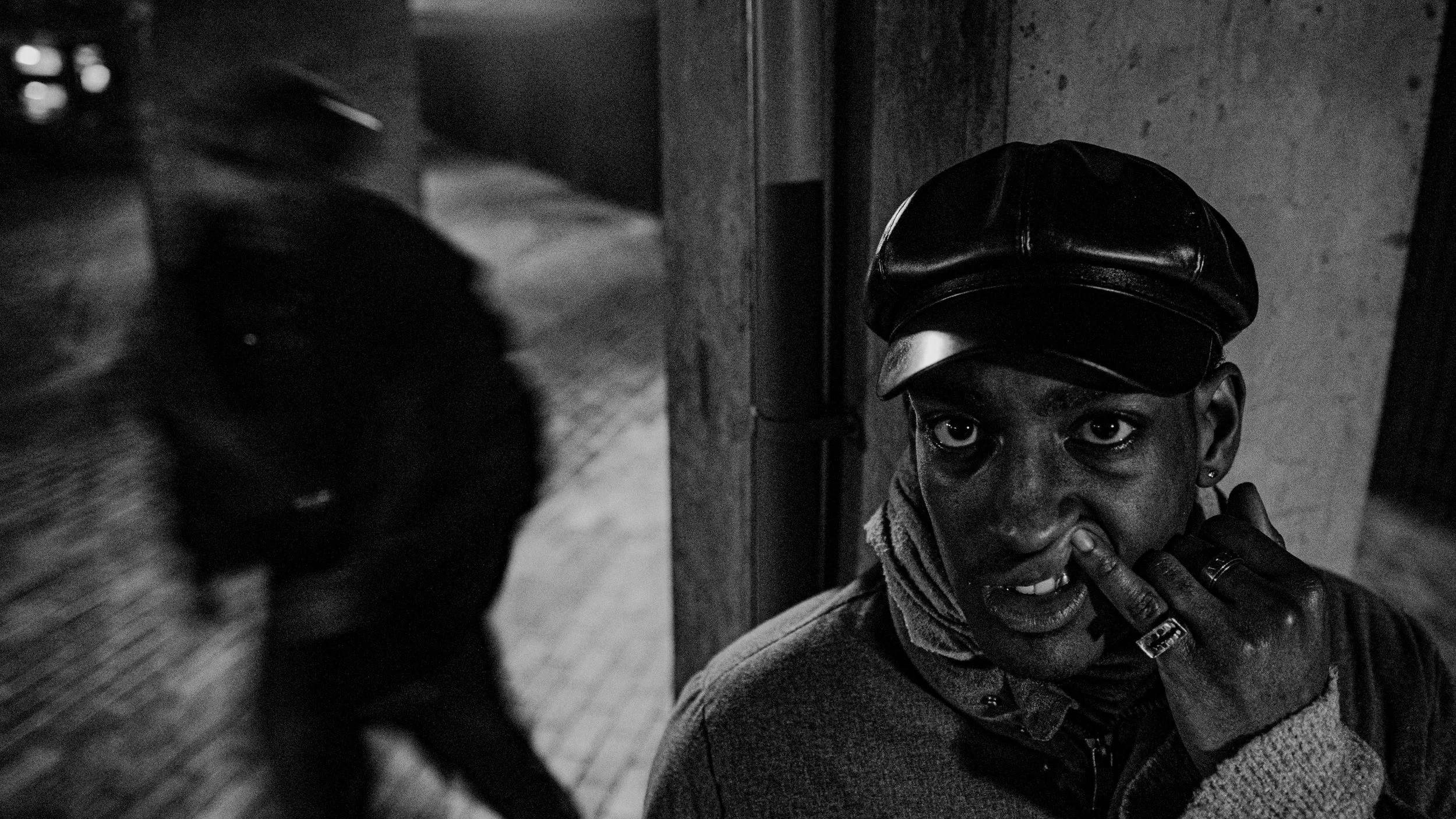 A young person with dark skin wearing a leather cap, holding fingers near mouth, and covering part of face in a black-and-white photo.