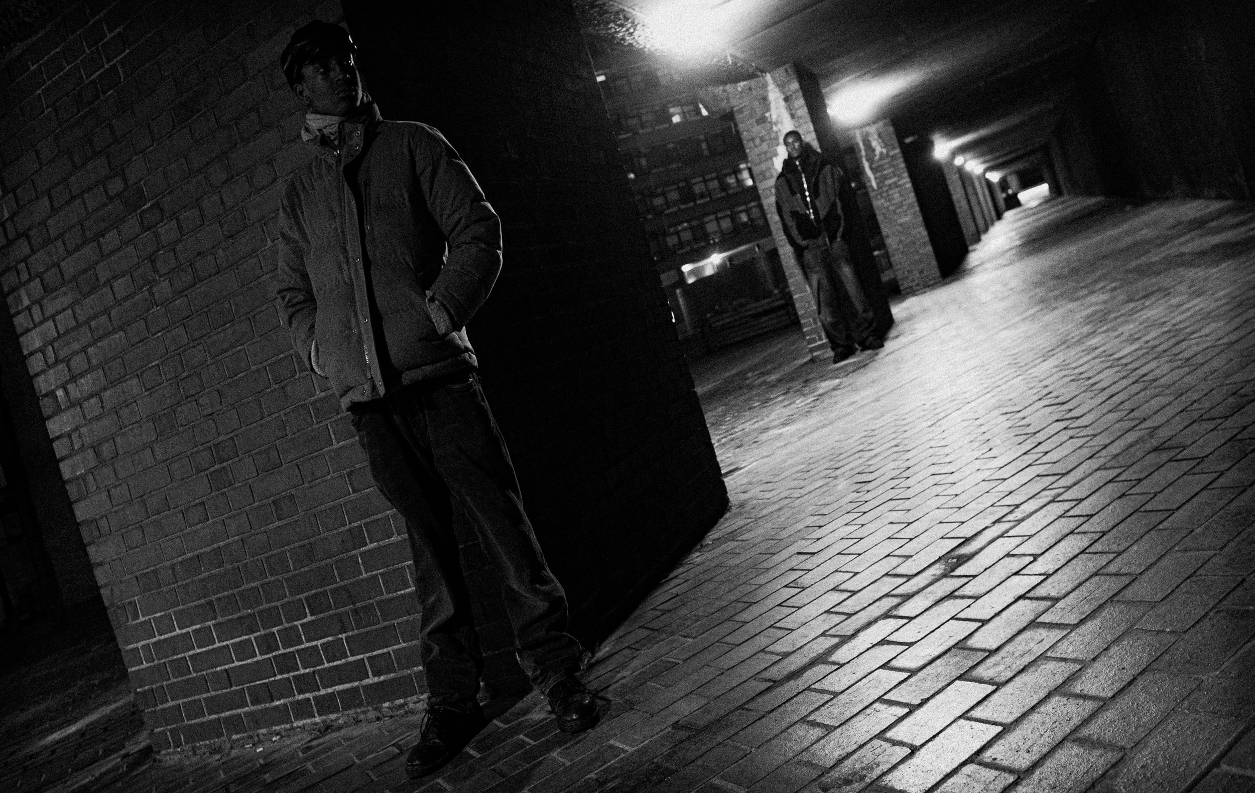 Two young men standing in a darkly lit alleyway at night. One is leaning against a brick wall with hands in pockets, and the other is standing further back near a building with a backpack. The scene is monochrome with streetlights illuminating the sc