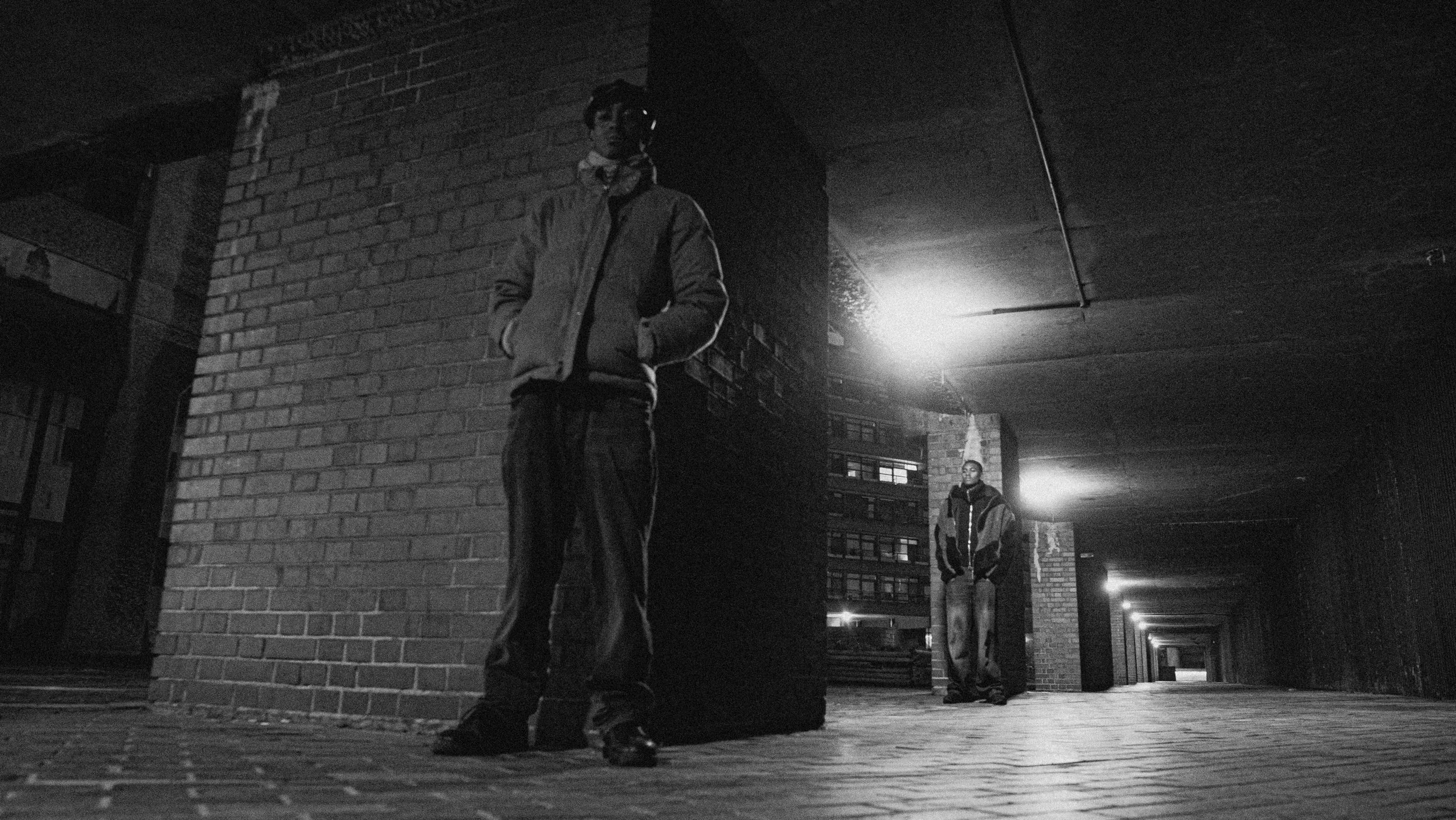 Two young men standing under a concrete overpass at night. The man in the foreground leans against a brick pillar with hands in his jacket pockets, wearing a hoodie and pants. The second man stands further in the background, also with hands in his po