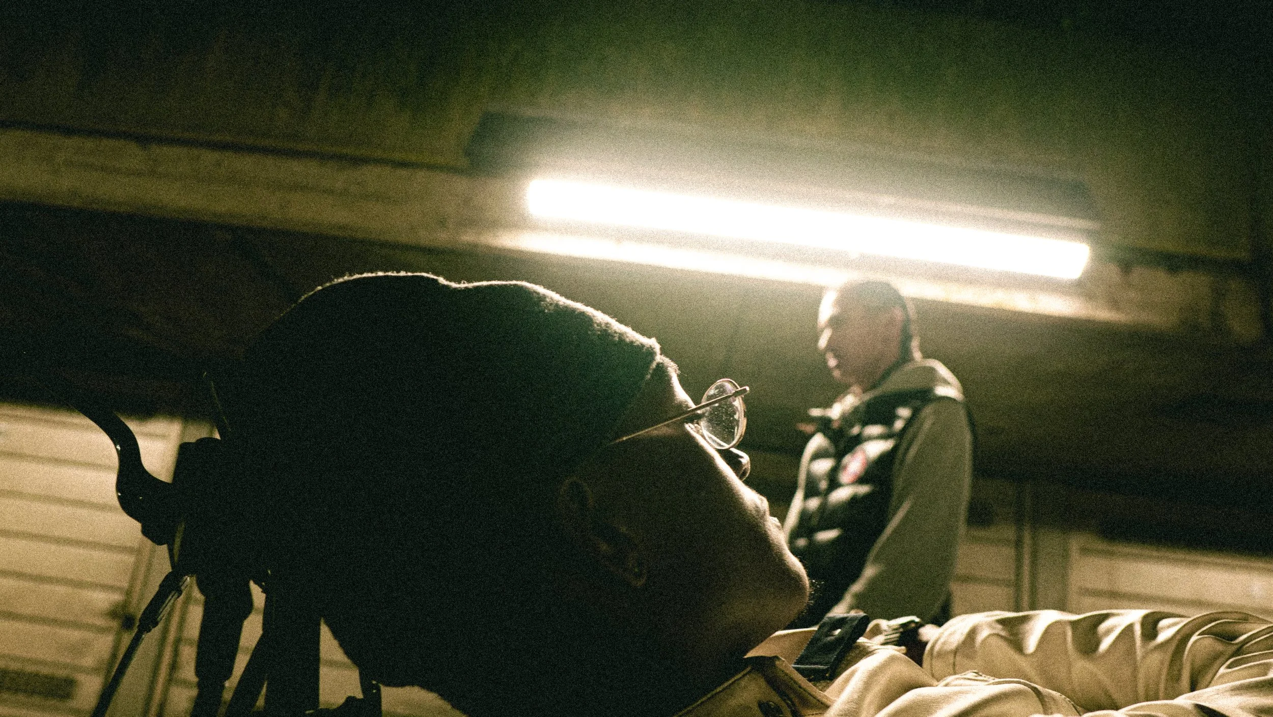 A person lying on a bed with a pair of glasses and a hat on their head, looking up at the illuminated ceiling. Another person wearing glasses is sitting in the background, looking towards the person on the bed. The setting appears to be a garage or b