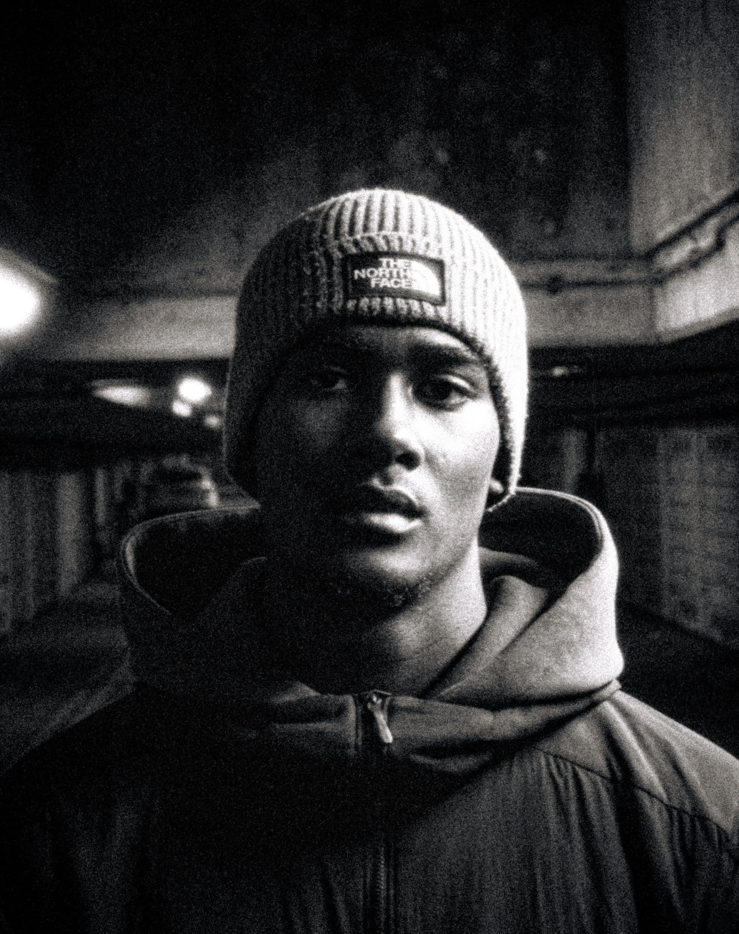 A black and white photo of a young man wearing a ribbed knit beanie with The North Face logo on it, a jacket with a zipper, and headphones around his neck. He is looking directly at the camera. The background appears to be indoors with some details l