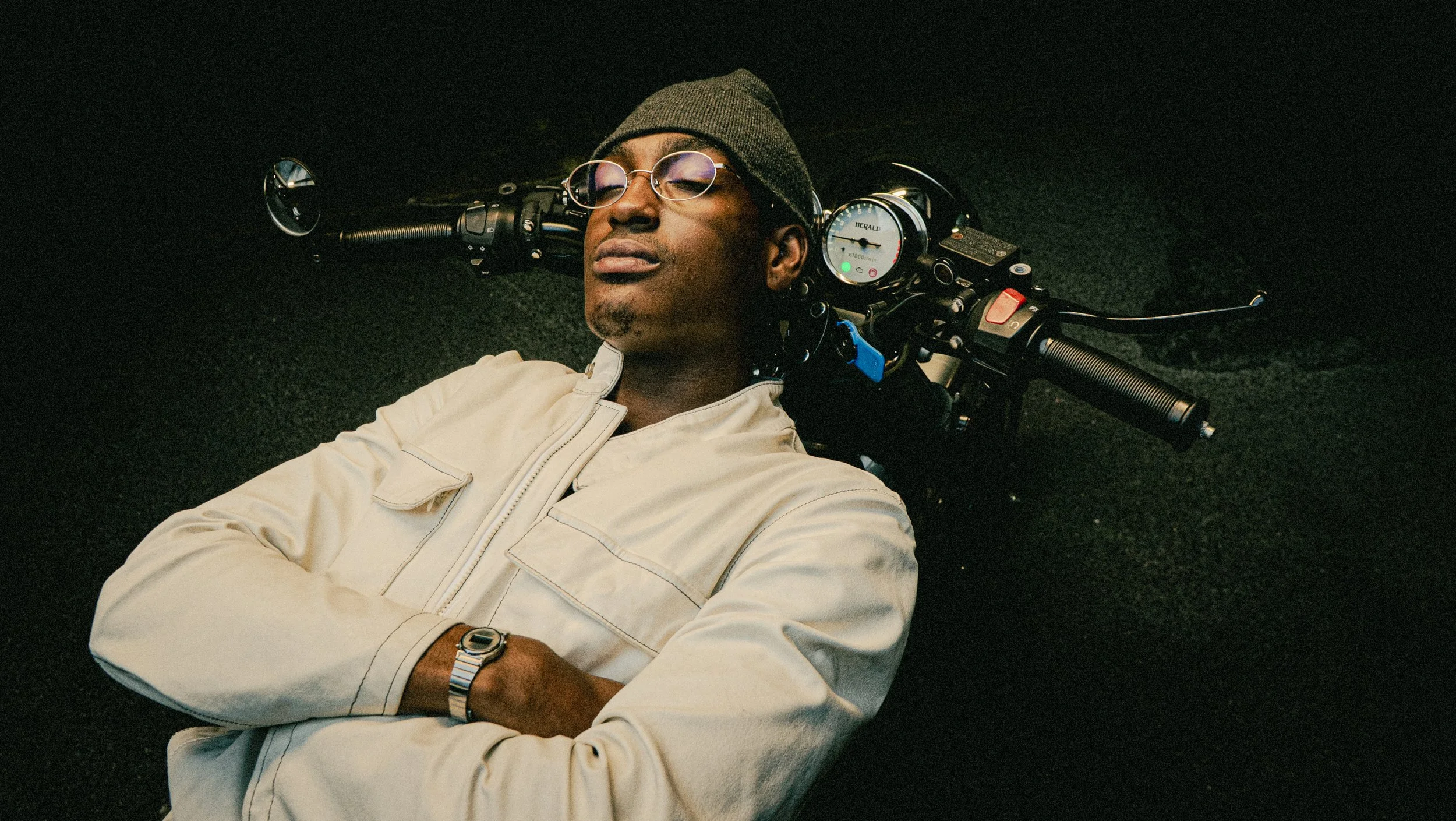A man with glasses and a beanie hat lying back on a motorcycle with a black background.