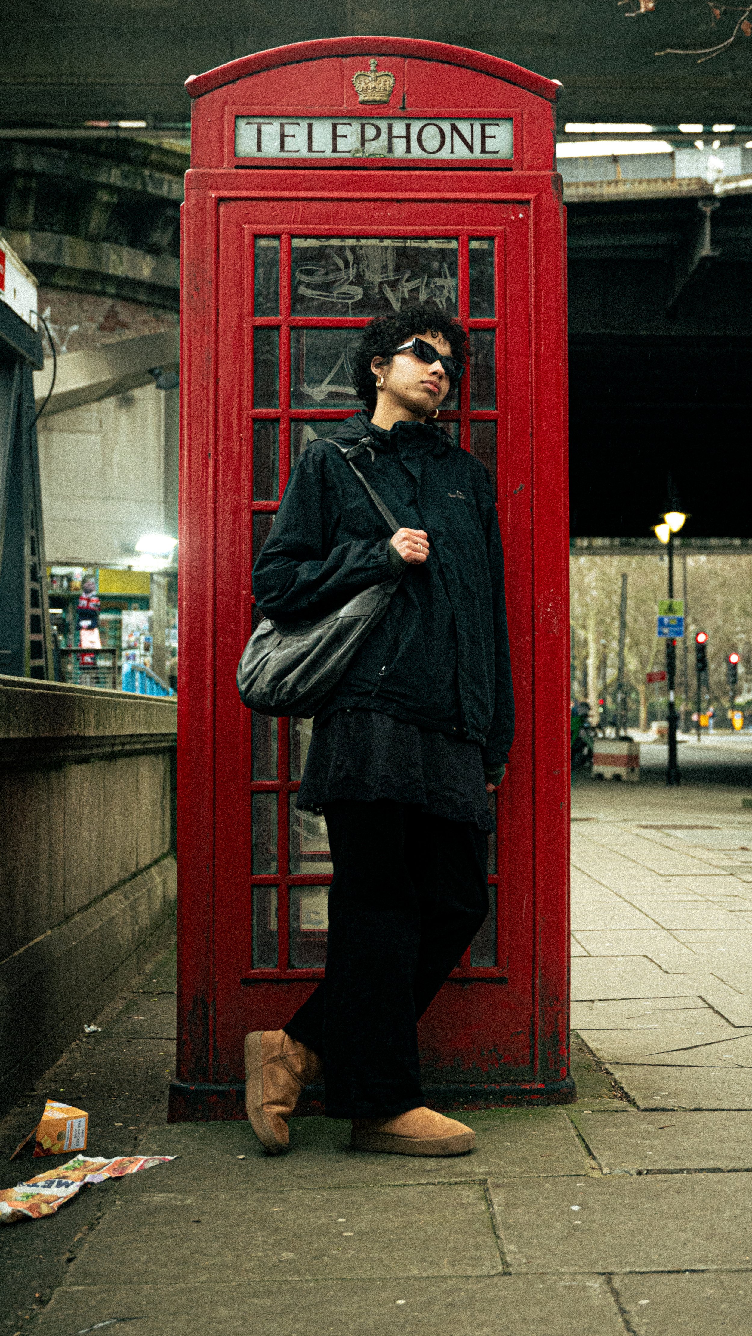 A woman with curly hair, black sunglasses, and gold hoop earrings standing in front of a red British telephone booth on a city sidewalk during the evening, wearing a black jacket, black pants, tan boots, and carrying a black shoulder bag.