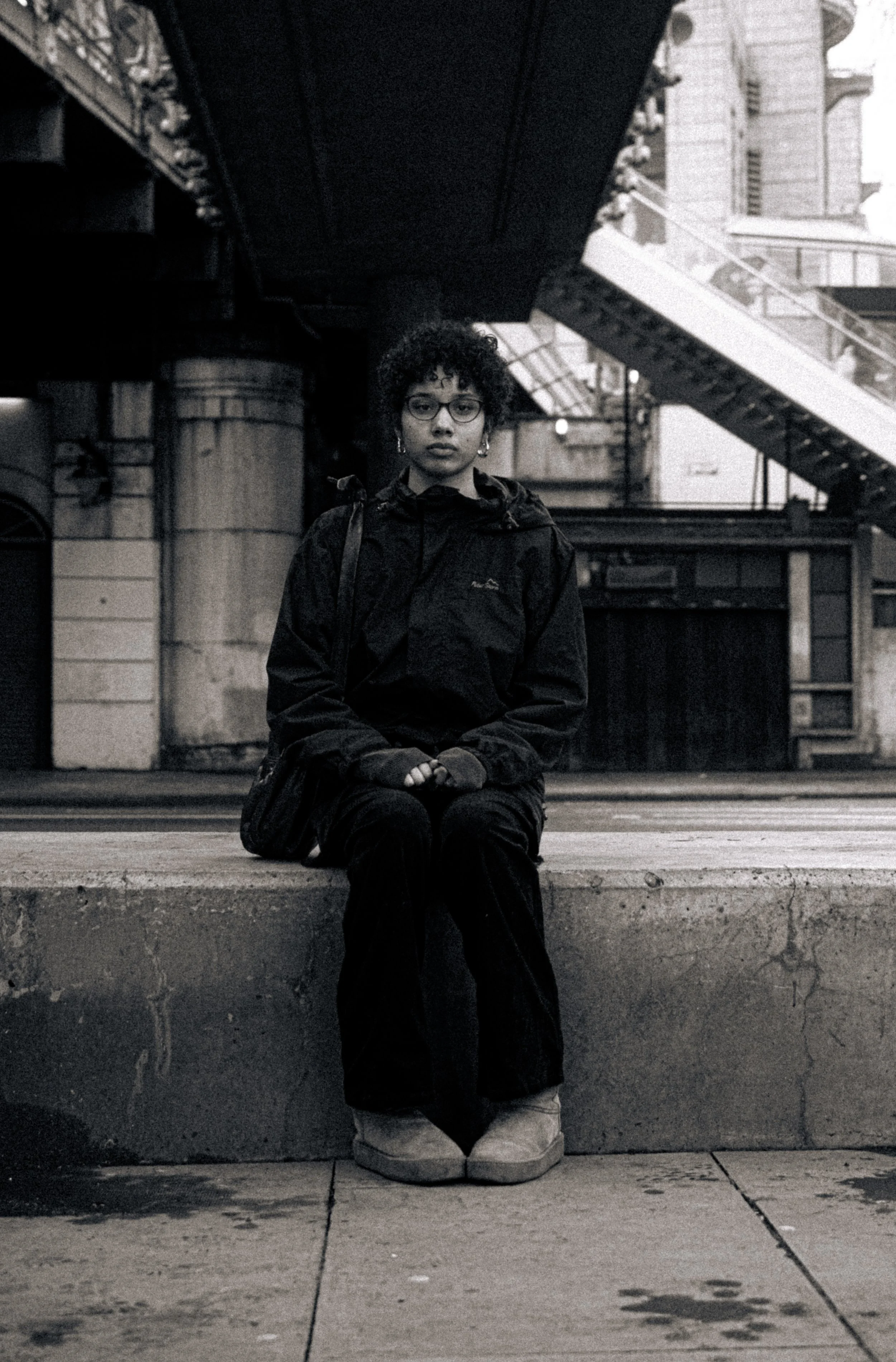 A young woman with curly hair, glasses, and earrings rides a skateboard while sitting on a sidewalk in an urban environment, with an overpass and buildings in the background, in black and white.