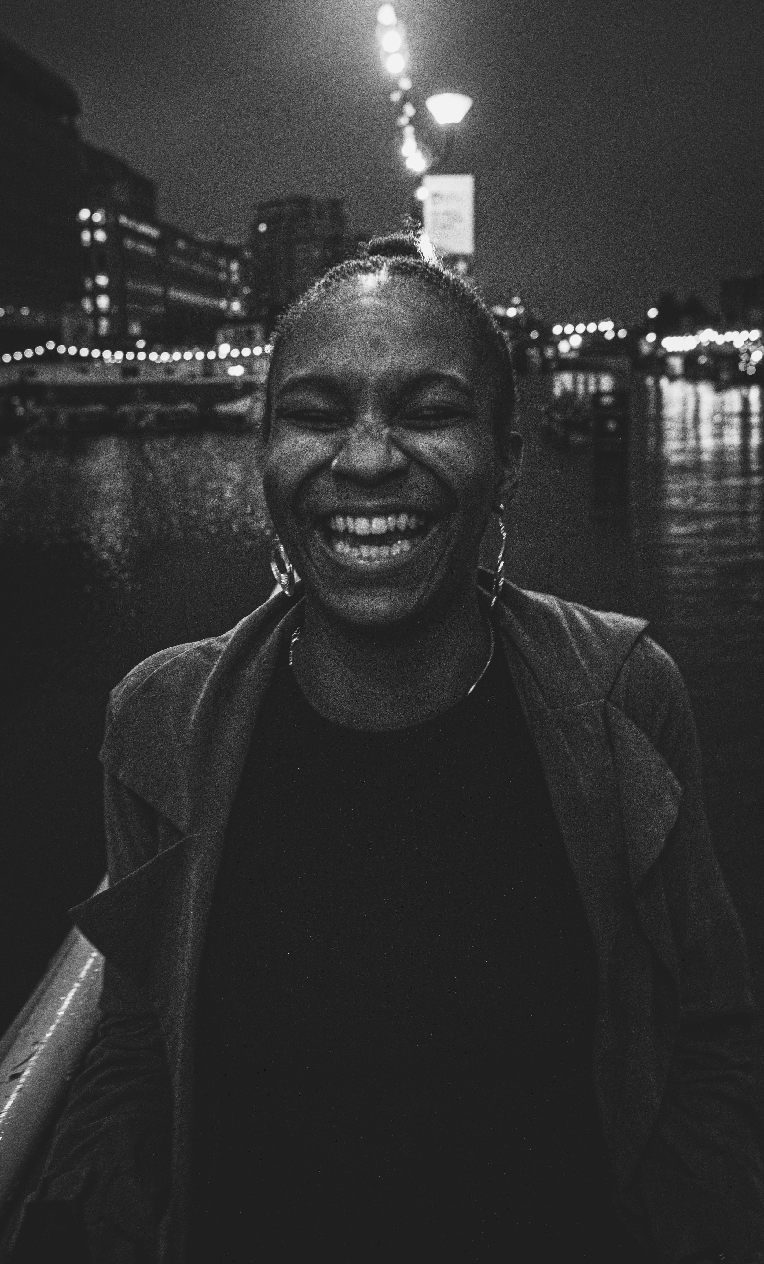 A young woman laughing on a rainy night by a waterfront with streetlights and buildings in the background.