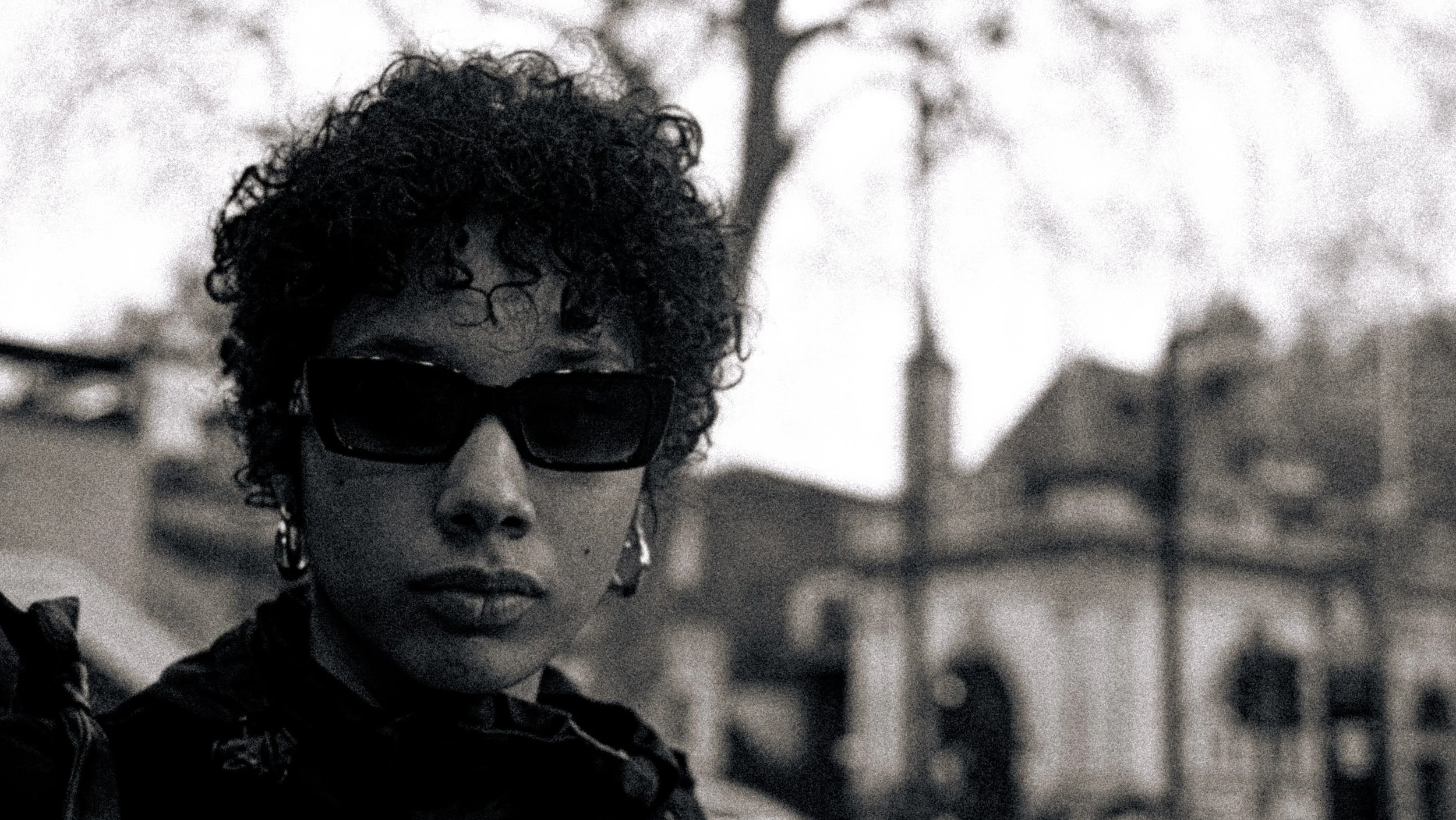 A young woman with curly hair, wearing sunglasses, outdoors with houses and trees in the background.