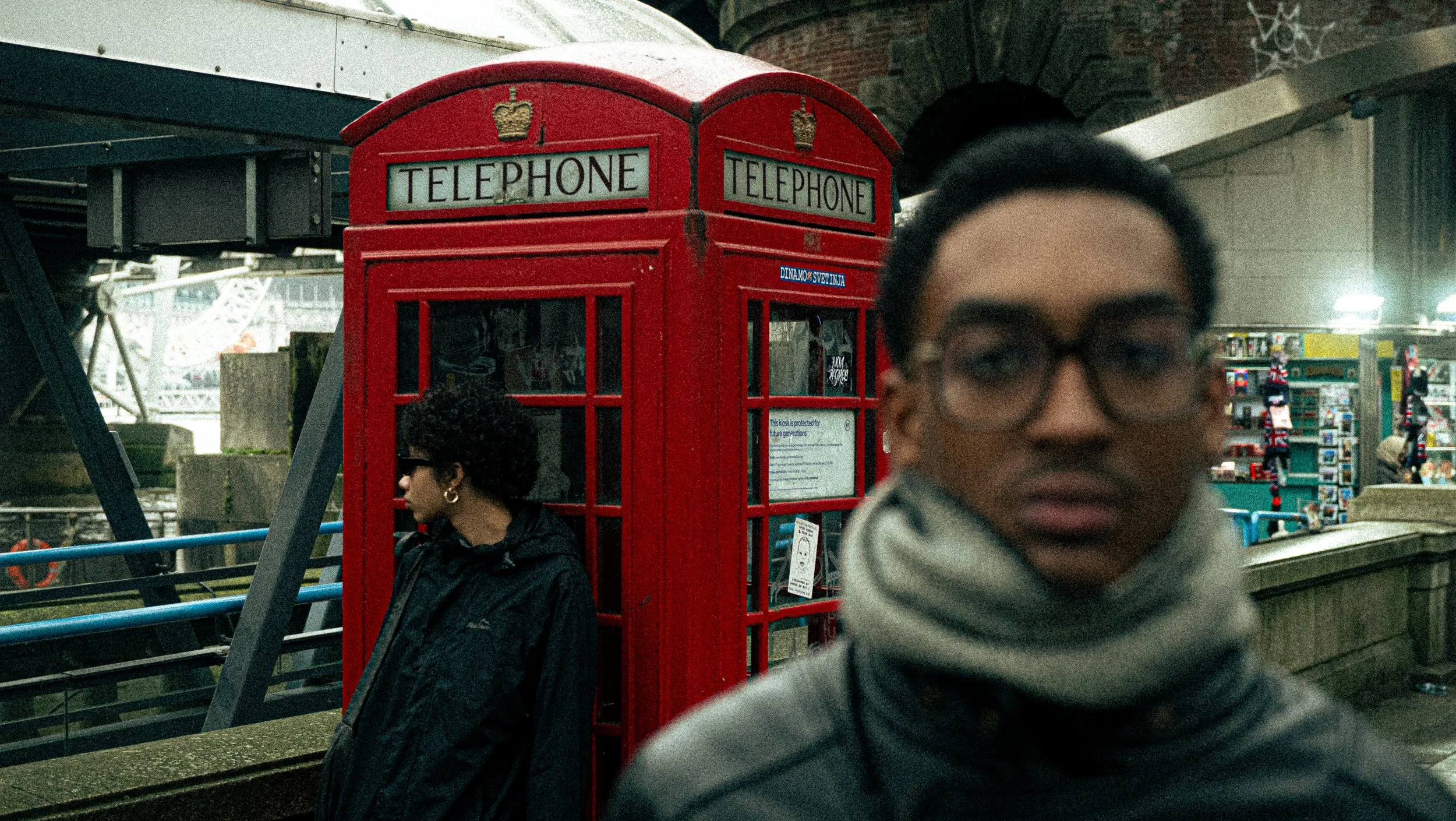 A red British telephone booth with calendar stickers on the side, positioned on a city sidewalk with two women nearby. One woman is in profile, wearing dark sunglasses and earrings, looking at her phone. The other woman is at the right edge, out of focus, with glasses and a striped scarf. A structure with an arched brick doorway and shop shelves can be seen in the background.