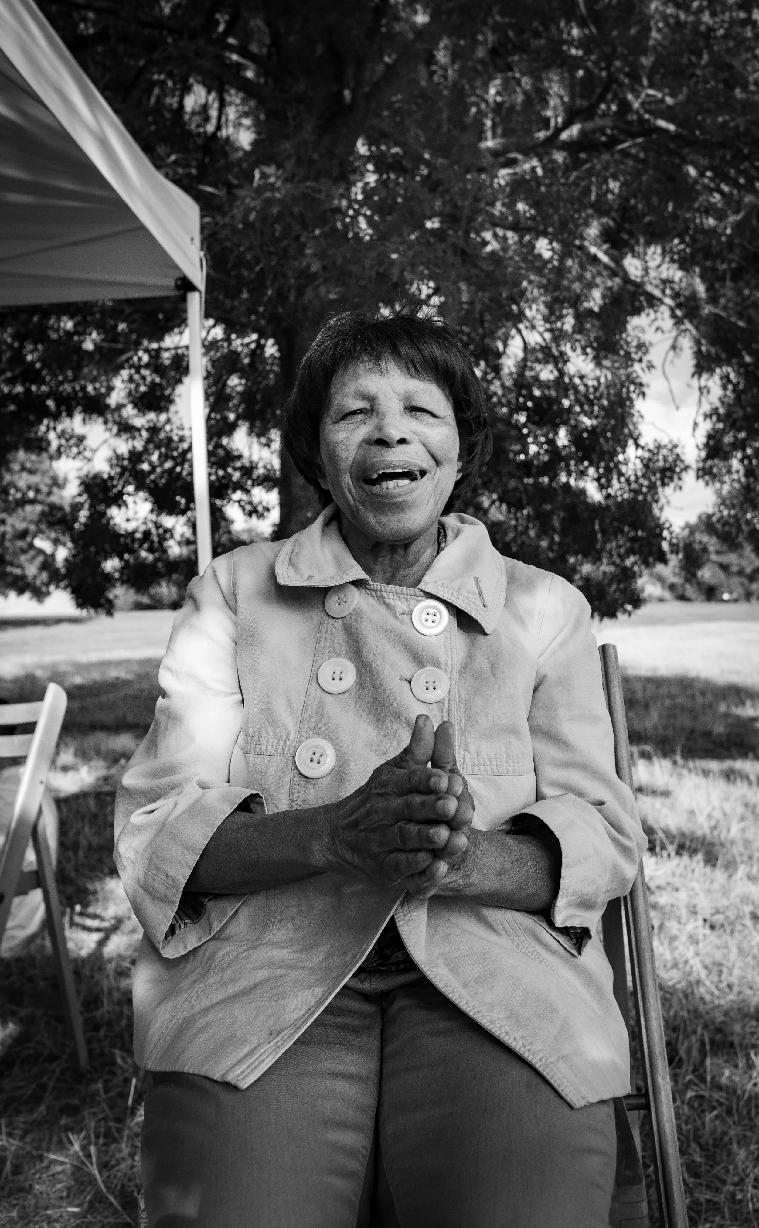 Black and white photo of a joyful woman sitting outdoors with hands clasped, under a large tree, with a canopy tent and chairs nearby.