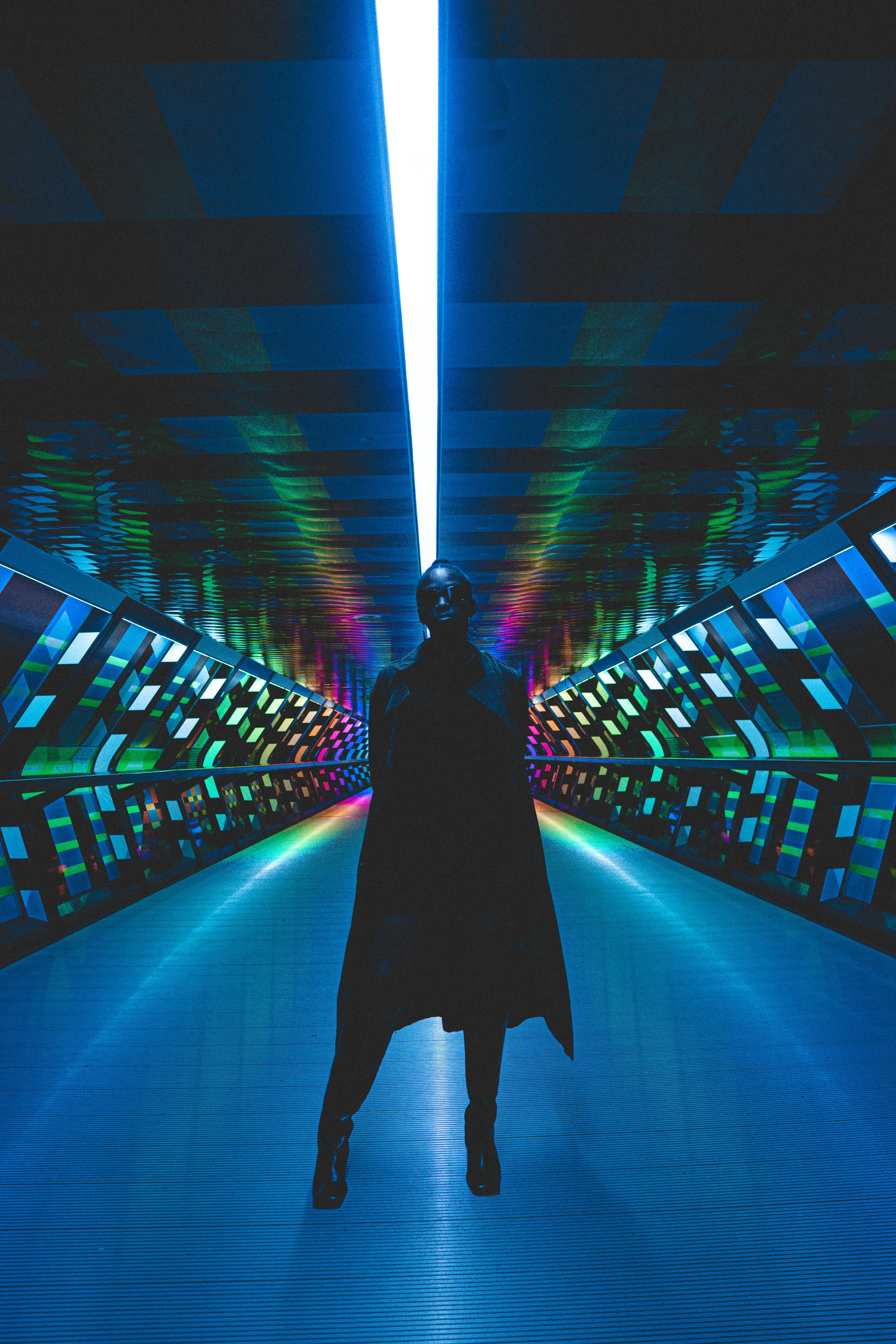 A silhouettes of a person standing in a colorful, futuristic corridor with blue, green, and purple lights and digital patterns on the ceiling and walls.