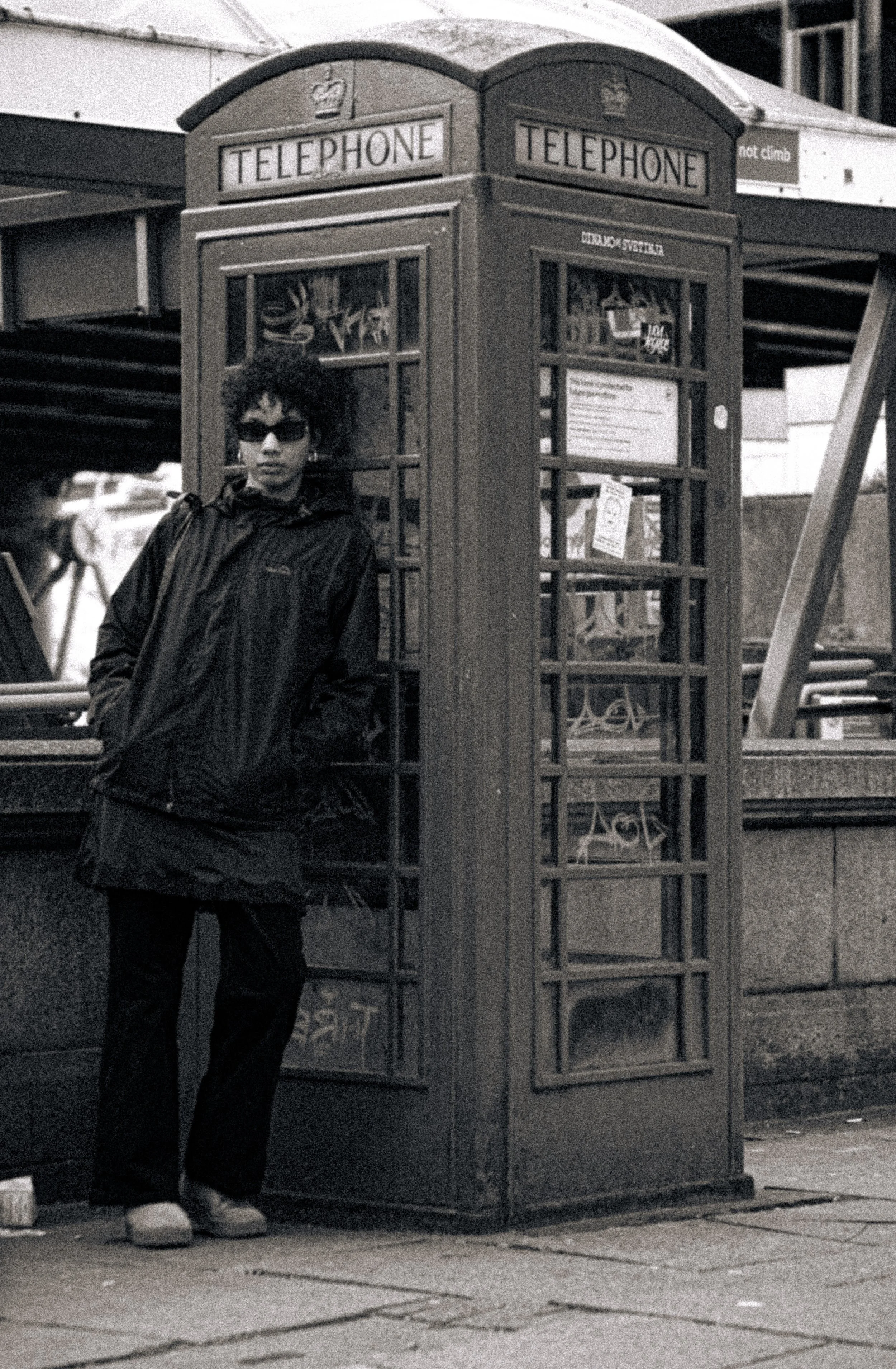 A person with curly hair and sunglasses standing next to a red telephone booth on a city street.