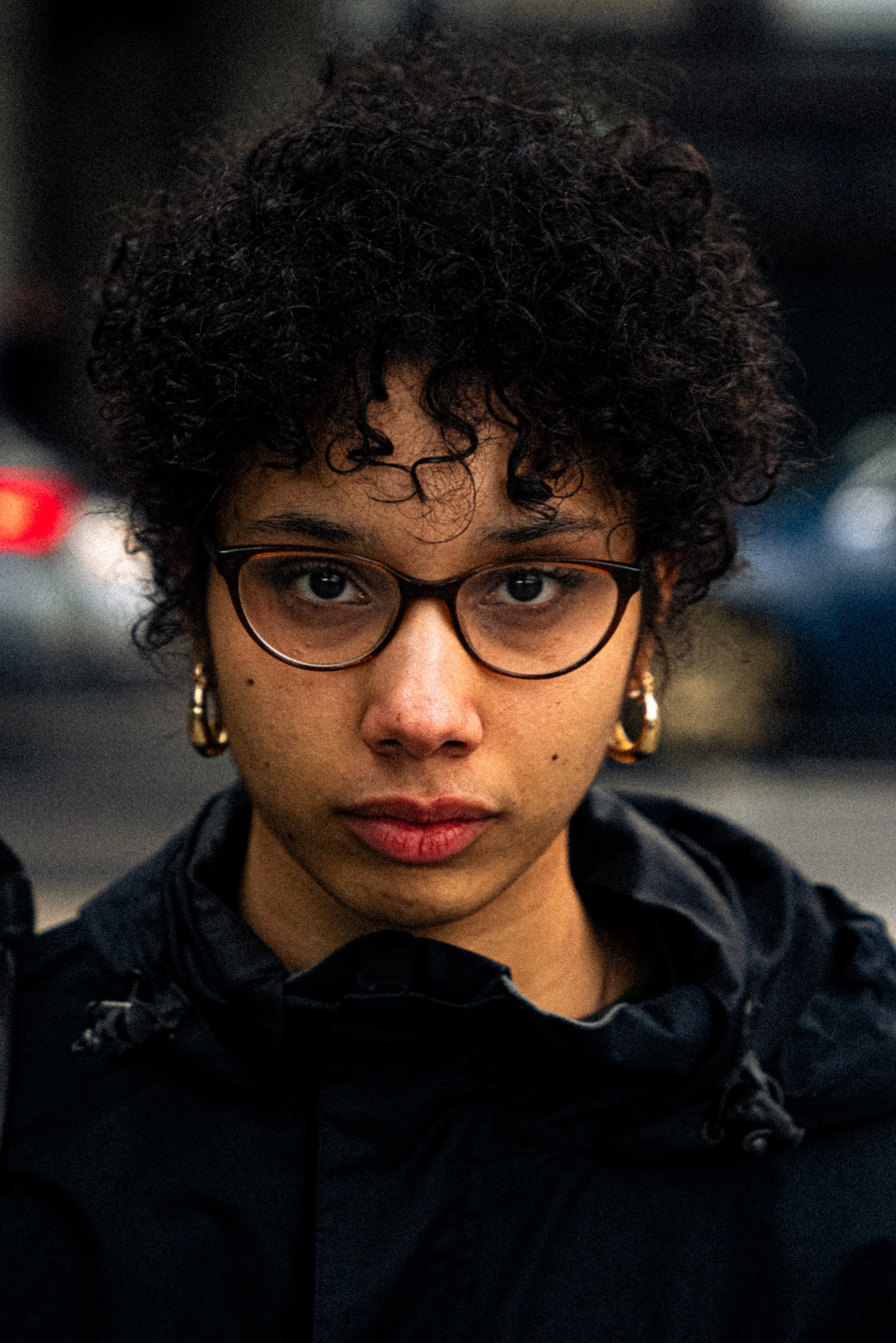 Close-up of a woman with short, curly dark hair, glasses, and earrings, wearing a black jacket in a dimly lit setting.