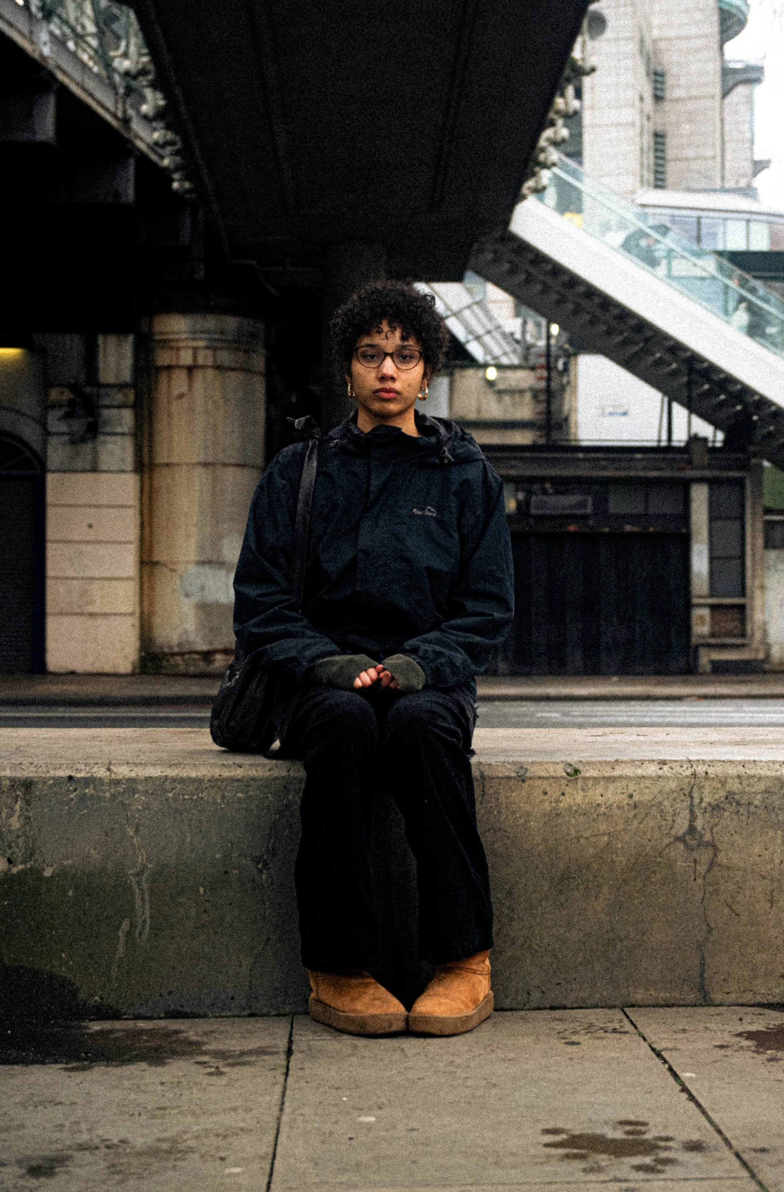 A young woman with curly hair and glasses sitting on a concrete curb under a bridge in an urban setting, wearing a dark jacket, black pants, and tan boots, with a backpack strap visible on her shoulder.