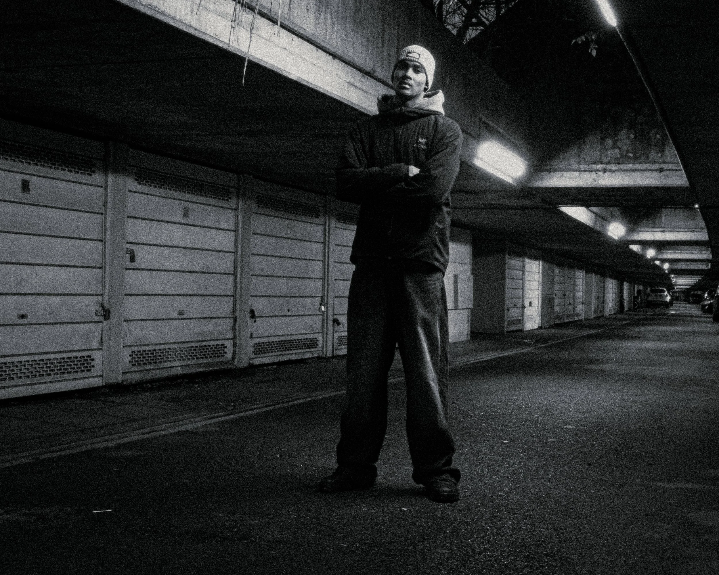 A person standing in a dark parking lot, wearing a beanie, jacket, and pants, with lockers and cars visible in the background.