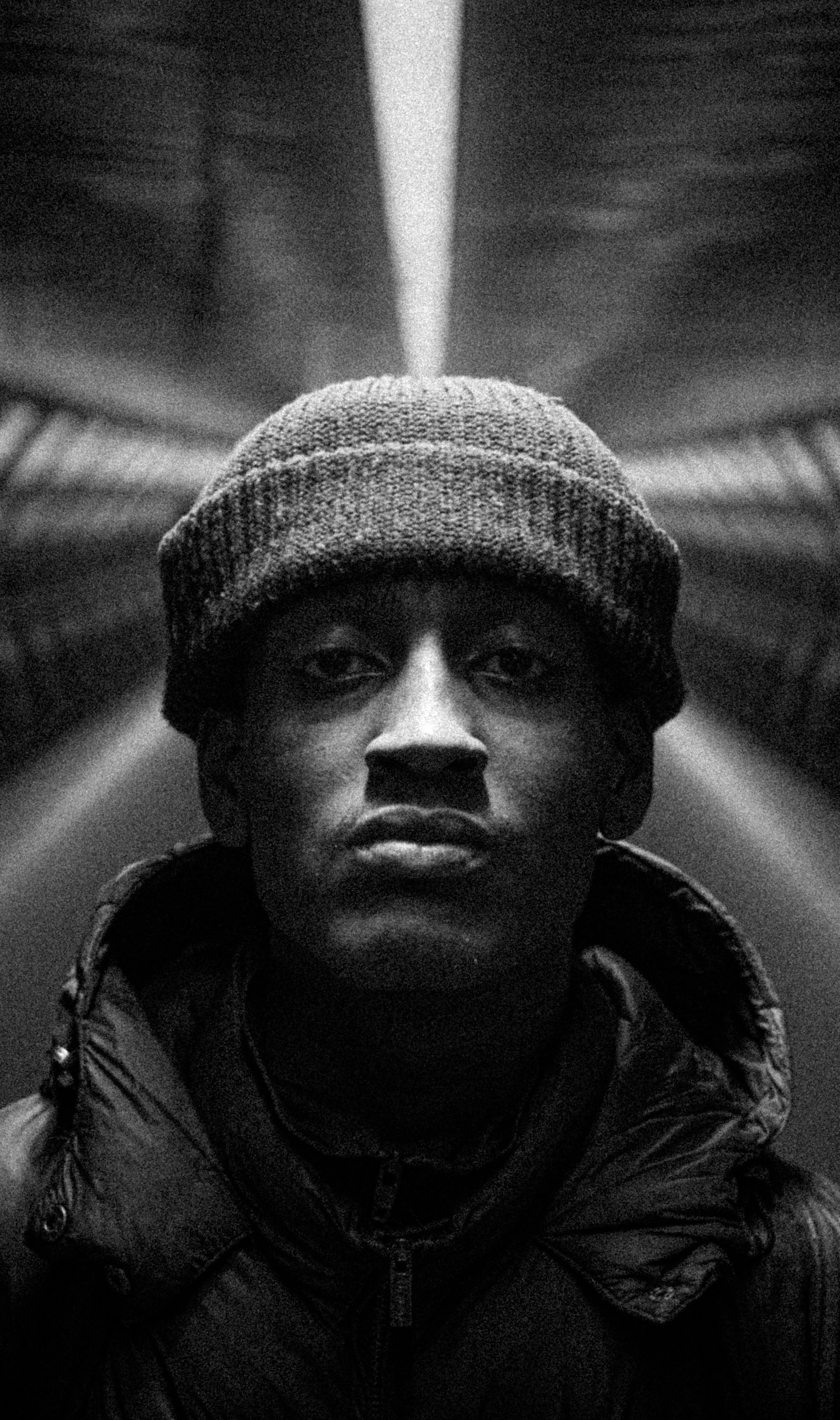 Black and white photo of a person wearing a knit beanie and a puffer jacket, looking into the camera with a serious expression.