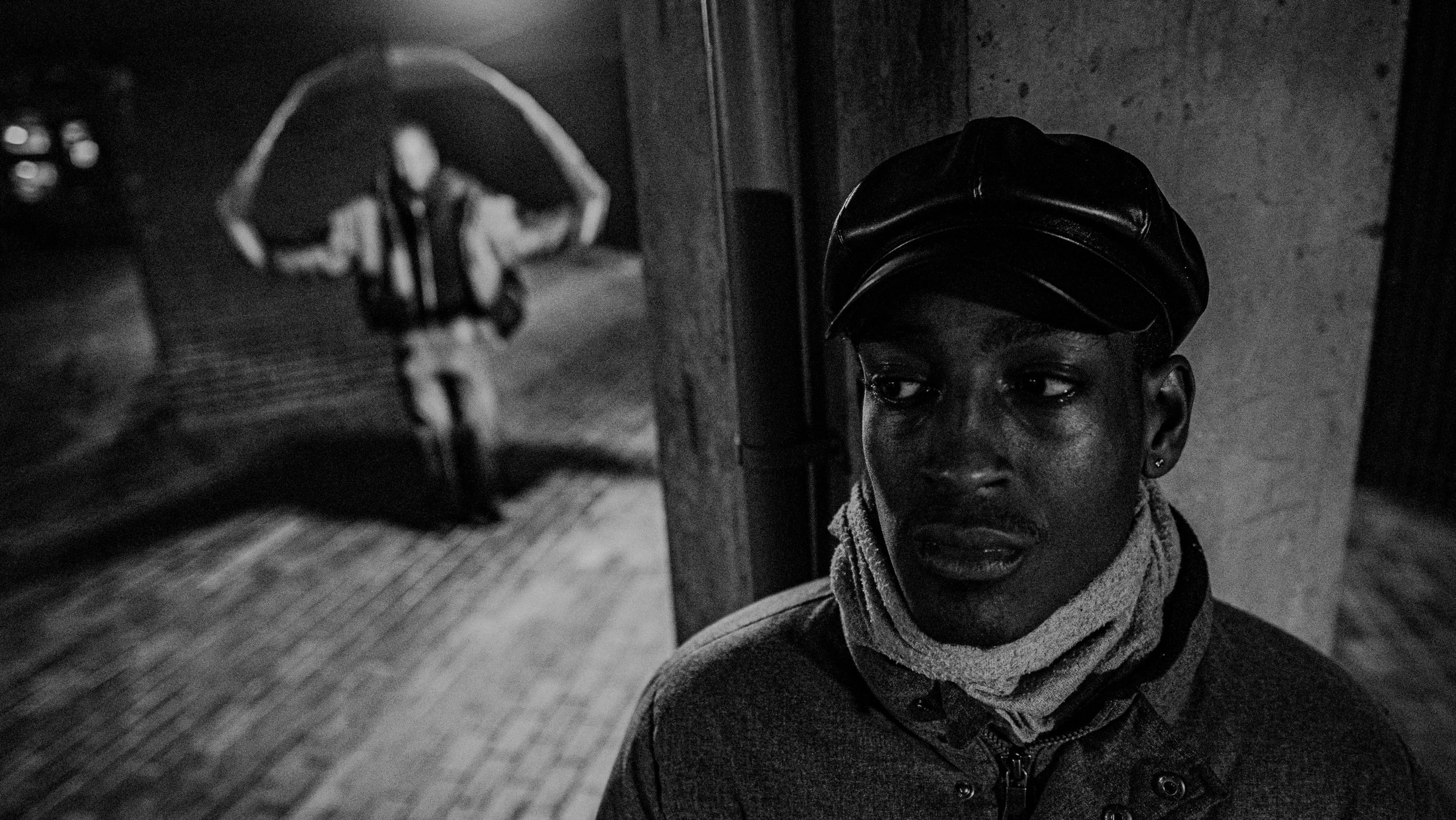 A Black boy wearing a cap and a scarf around his neck, looking to his side; reflected in a mirror.