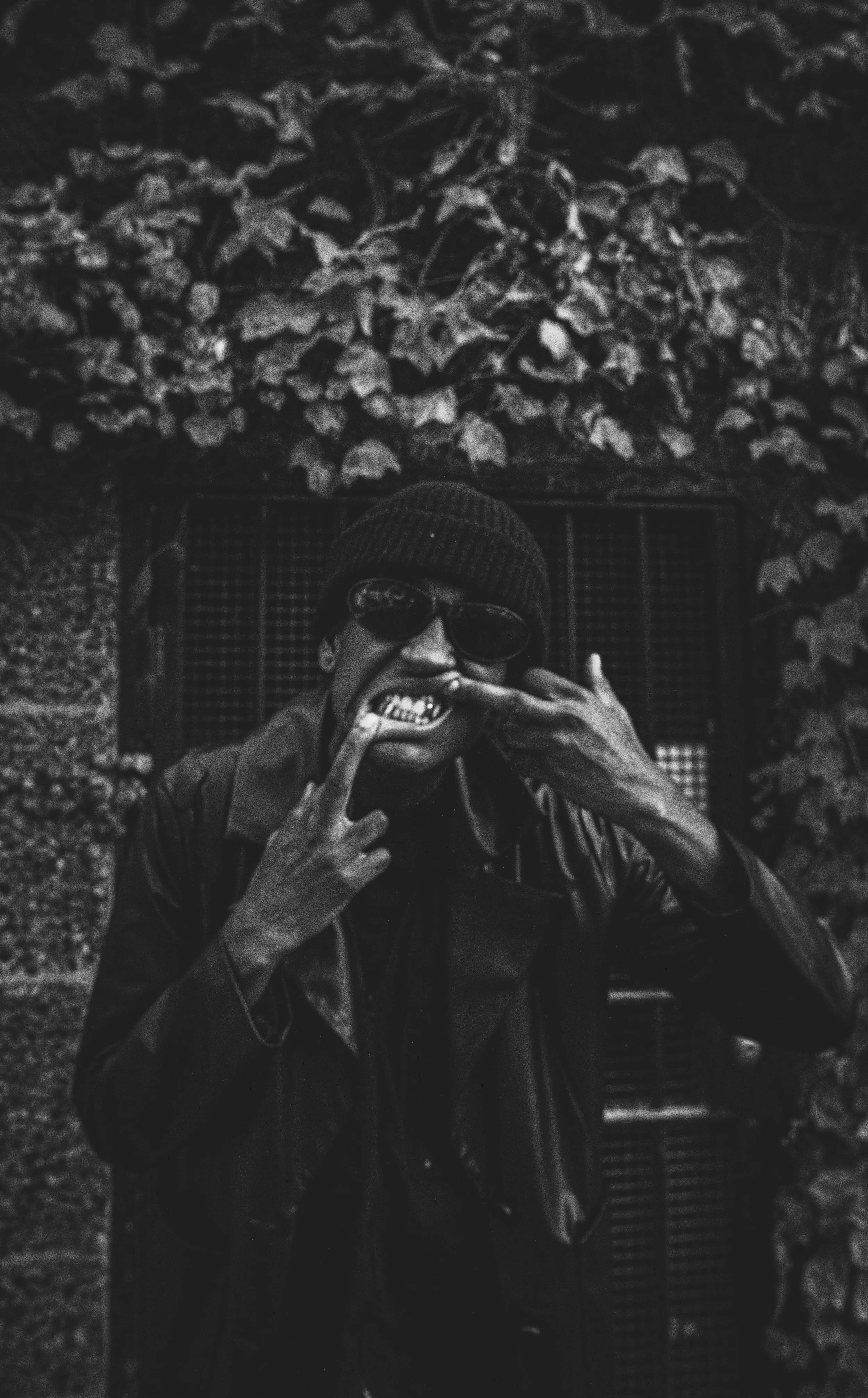 A person wearing a beanie and sunglasses taking a selfie in front of a wall with leafy vines, dressed in a dark leather jacket, making gestures with their hands.