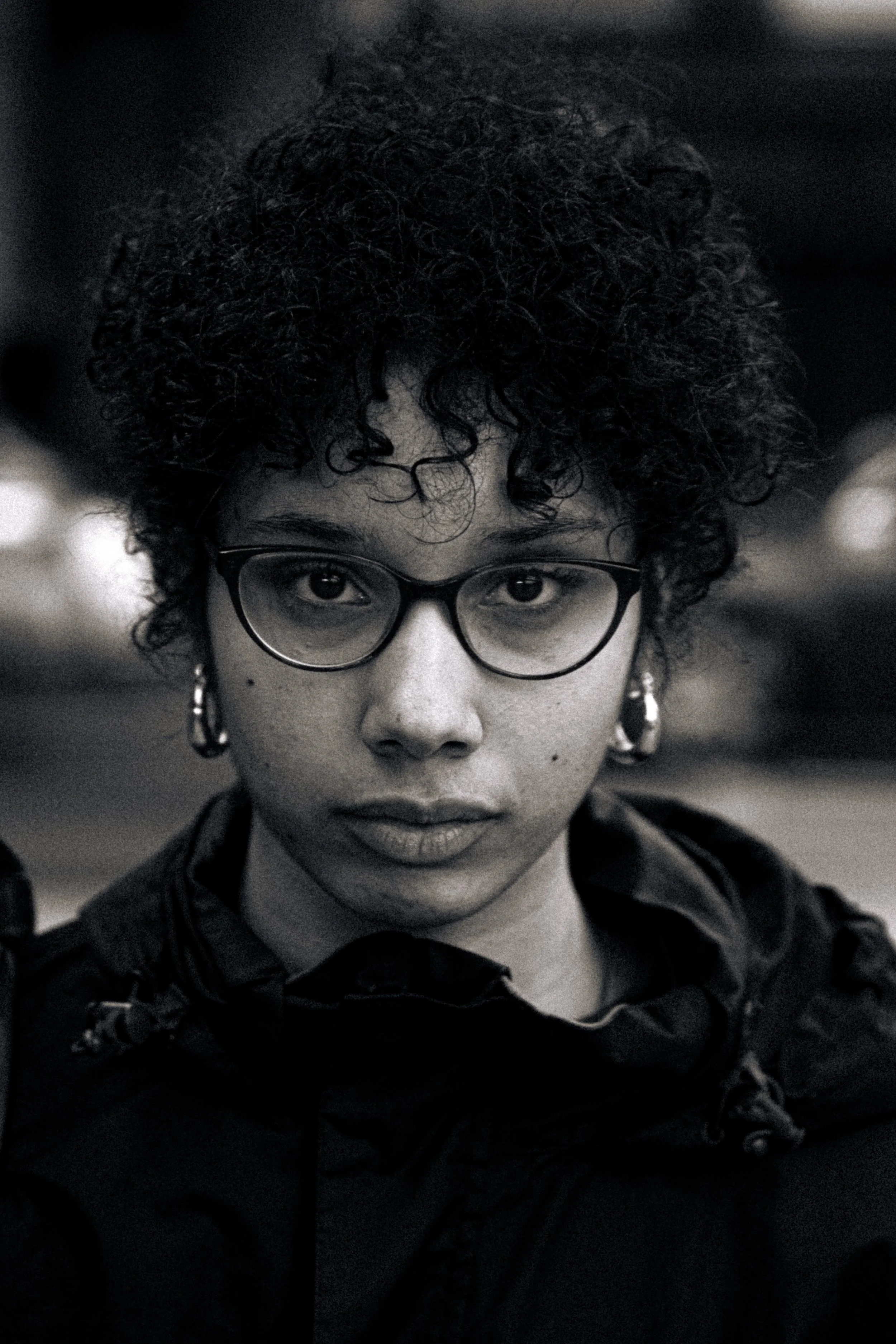 Black and white close-up portrait of a woman with curly hair, wearing glasses and earrings, looking directly at the camera.