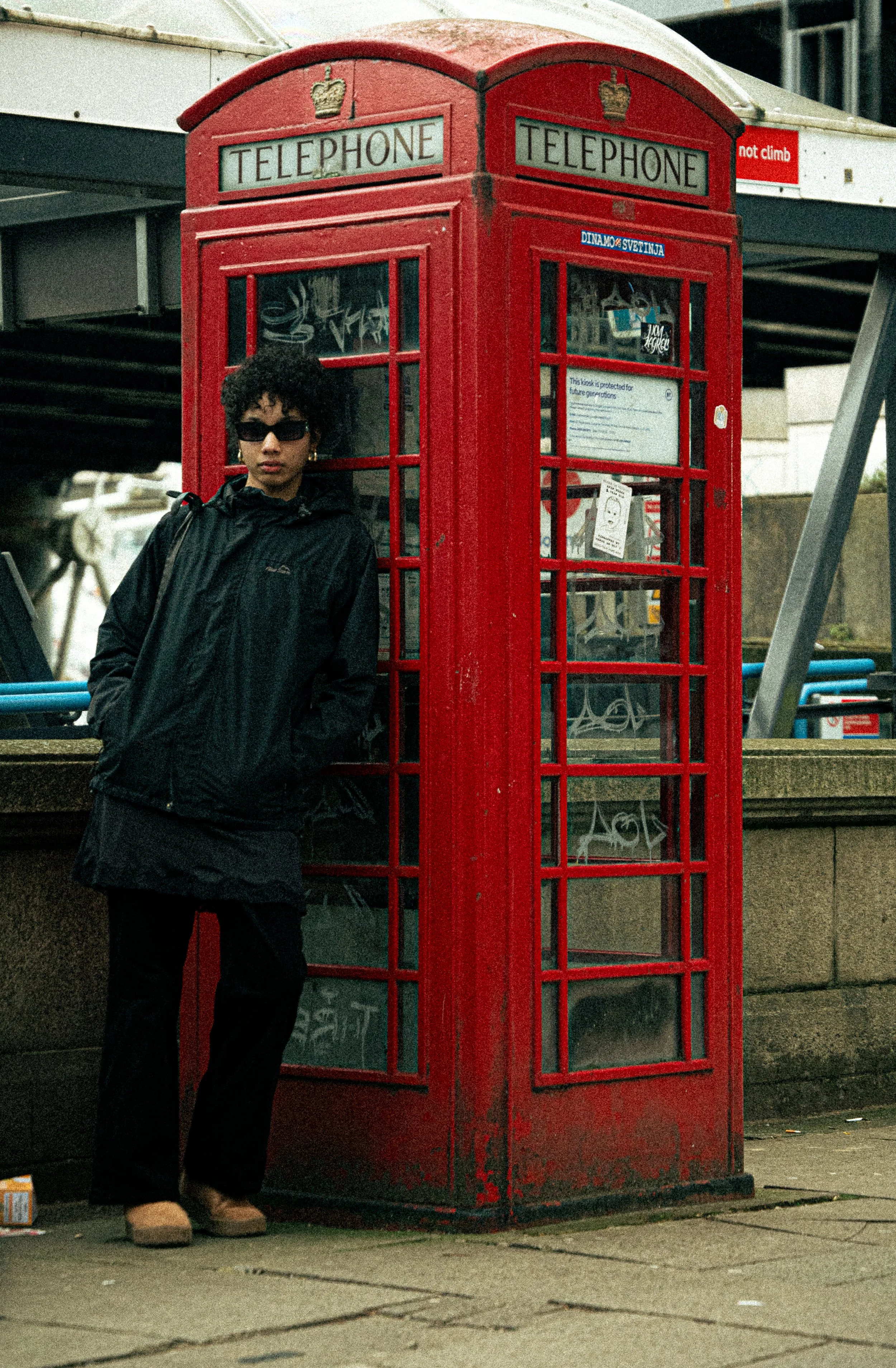 A person wearing black sunglasses, a black jacket, and beige boots stands beside a classic red British telephone booth on a city street. The booth has signs that say "Telephone" and a small sticker that says "not climb."