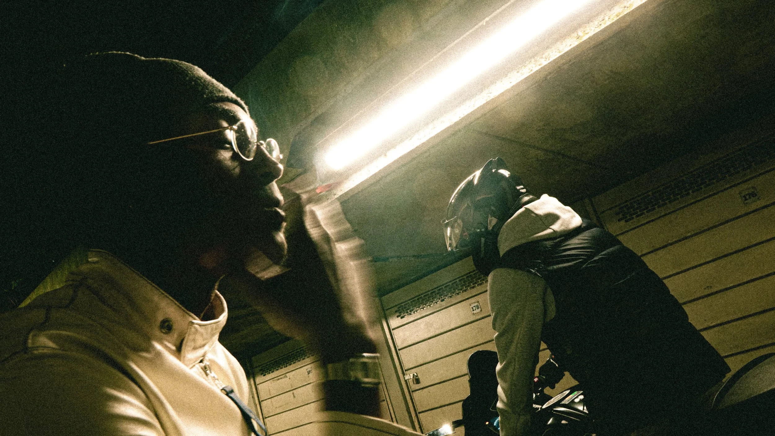 Two individuals wearing jackets, hats, and glasses are seated in an indoor parking garage near overhead doors and a wall with numbered lockers.