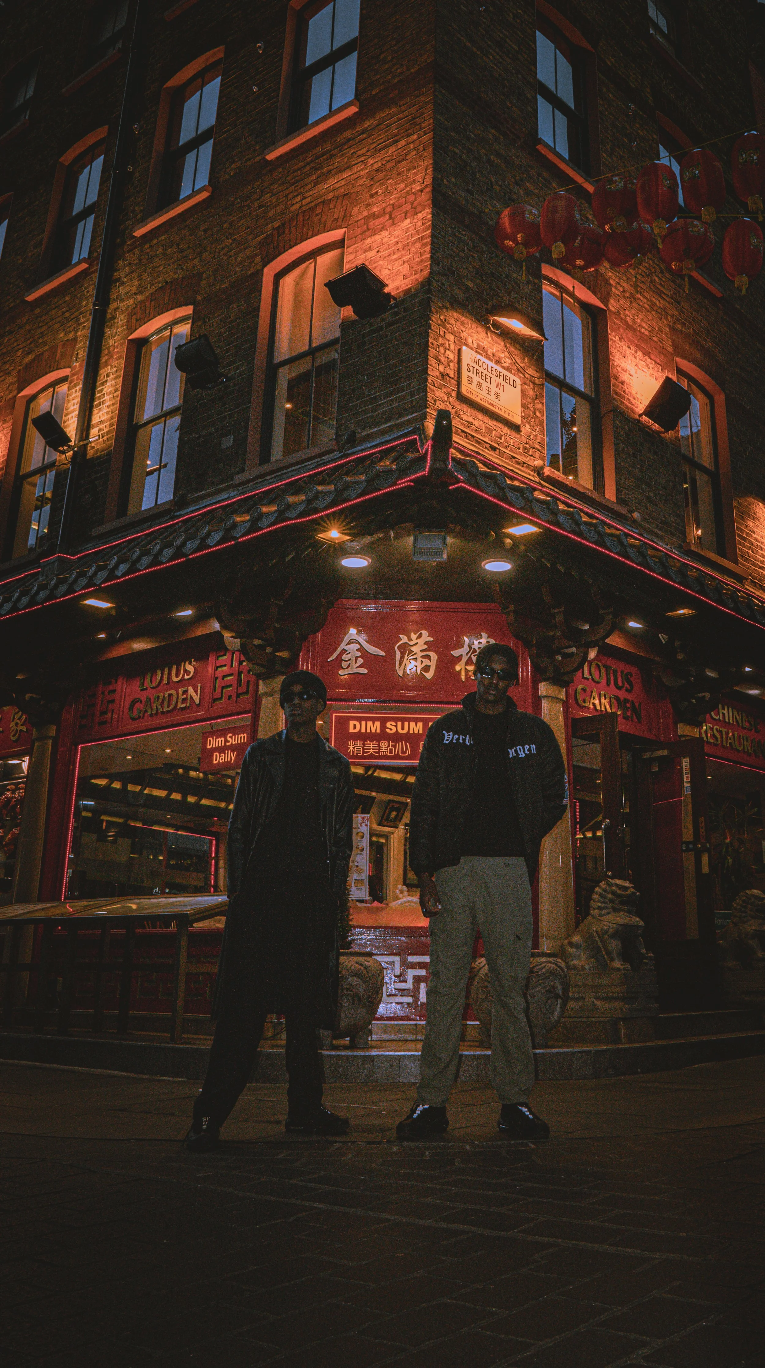 Two men standing in front of a Chinese restaurant called Lotus Garden on a street corner at night. The building has brick walls, multiple windows, and red lanterns hanging outside. The restaurant's entrance is lit with neon lights.