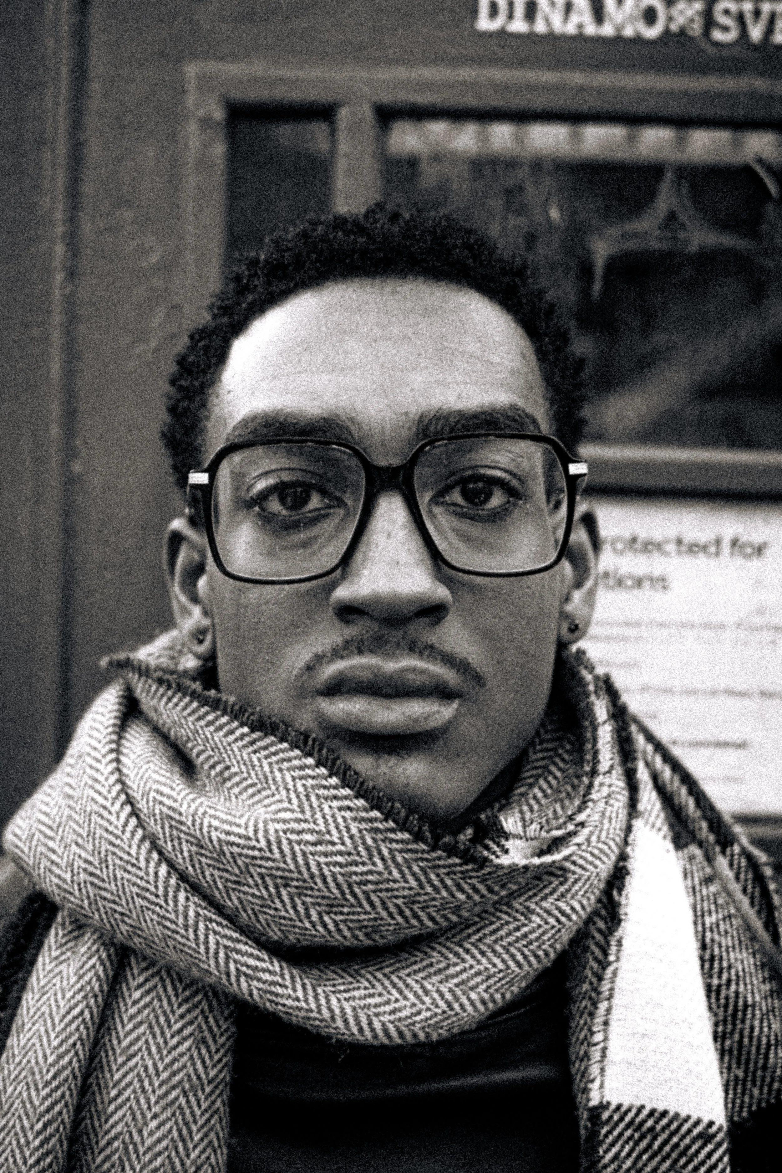 Close-up black and white photograph of a person with short curly hair, wearing glasses, a scarf with a herringbone pattern, and earrings, with signs and a window or display in the background.