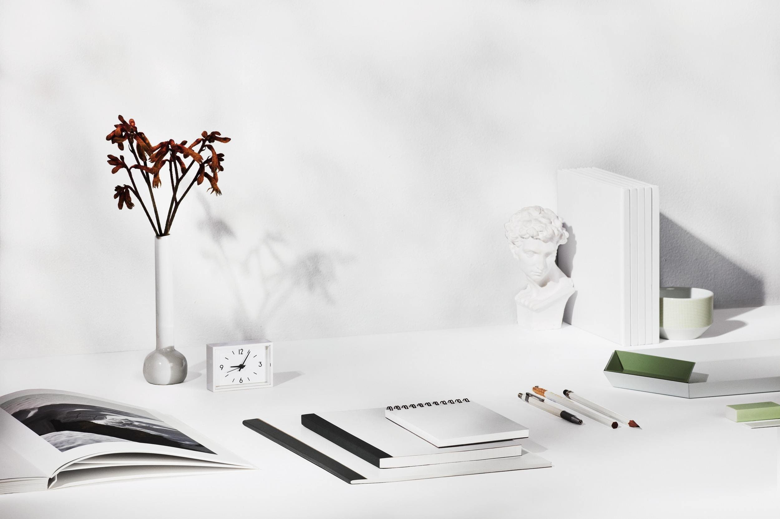 Minimalist white desk with dried flowers in a tall white vase, a small alarm clock, open book, notebooks, pens, a green folder, a green and white bowl, a few white books, and a white sculpture of a classical head against a plain white wall.