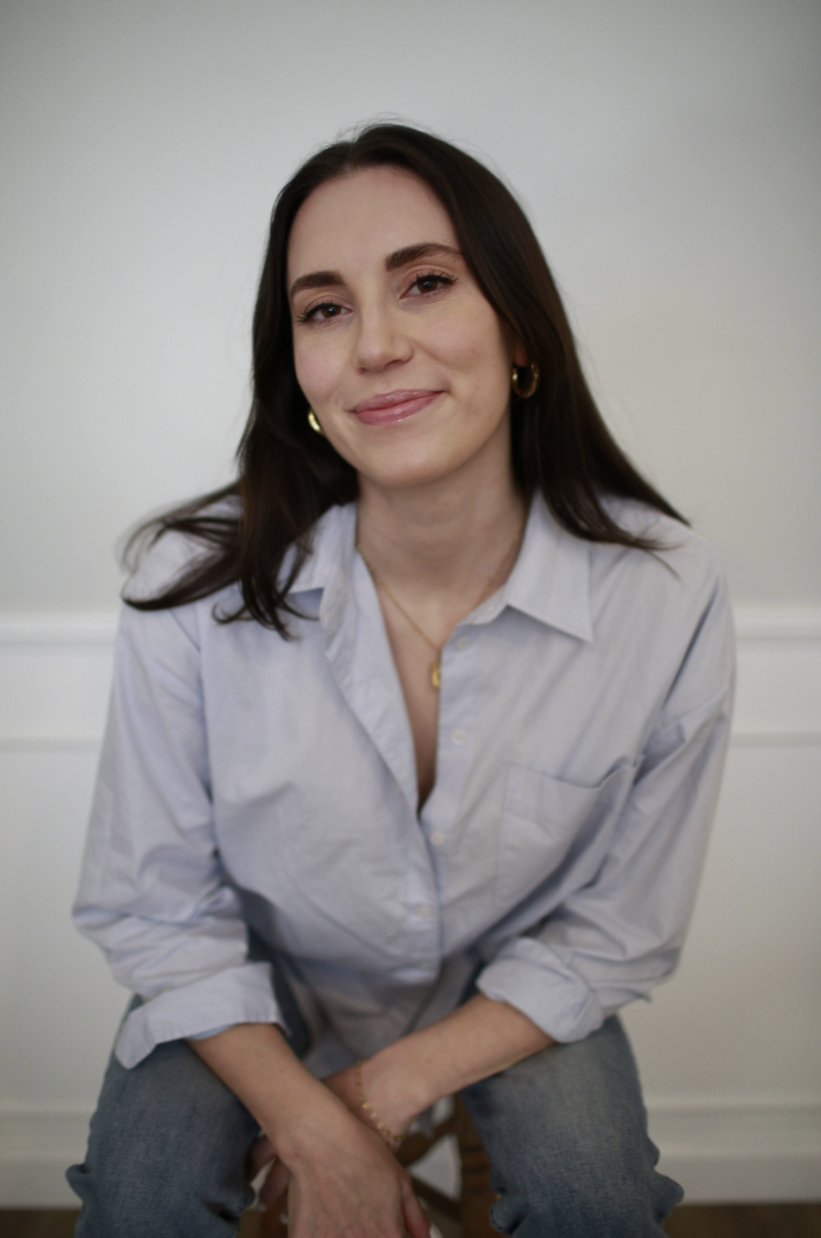 A woman with dark brown hair and fair skin, wearing a light blue button-up shirt and gold jewelry, smiling and looking at the camera against a plain light-colored background.