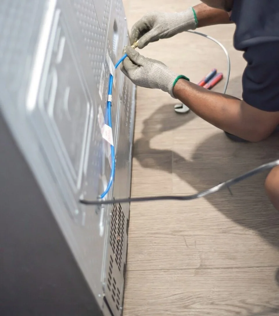 A person wearing gloves and a dark shirt is installing or repairing electrical wiring in the back of a metallic electrical panel or appliance, with wiring tools nearby.