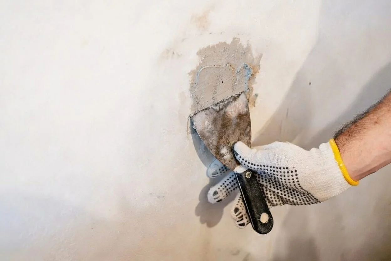 Person in a work glove using a putty knife to scrape old paint or drywall from a wall during repair or renovation.