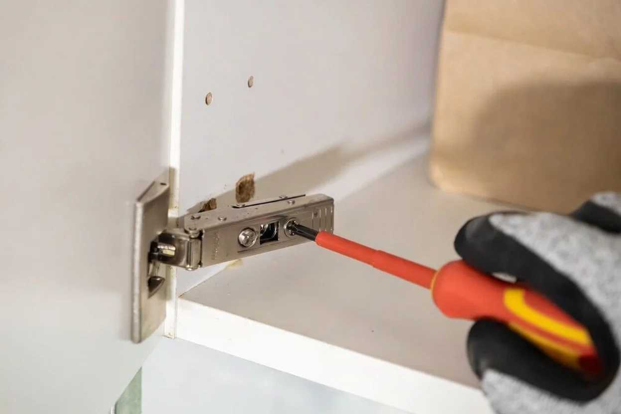 A person's hand wearing black and white work gloves using a screwdriver to install or repair a cabinet hinge inside a white cabinet door.
