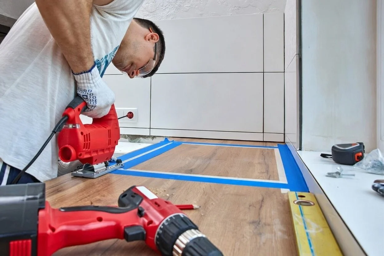 A person is using a power jigsaw to cut a wooden floor panel while installing flooring in a room. Blue painter's tape outlines the cutting area and tools are on the floor.