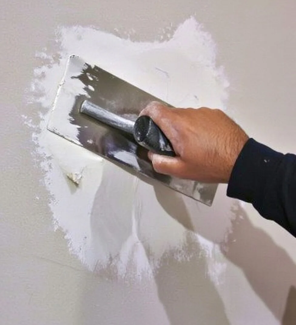Person using a trowel to apply joint compound around a wall electrical box opening.