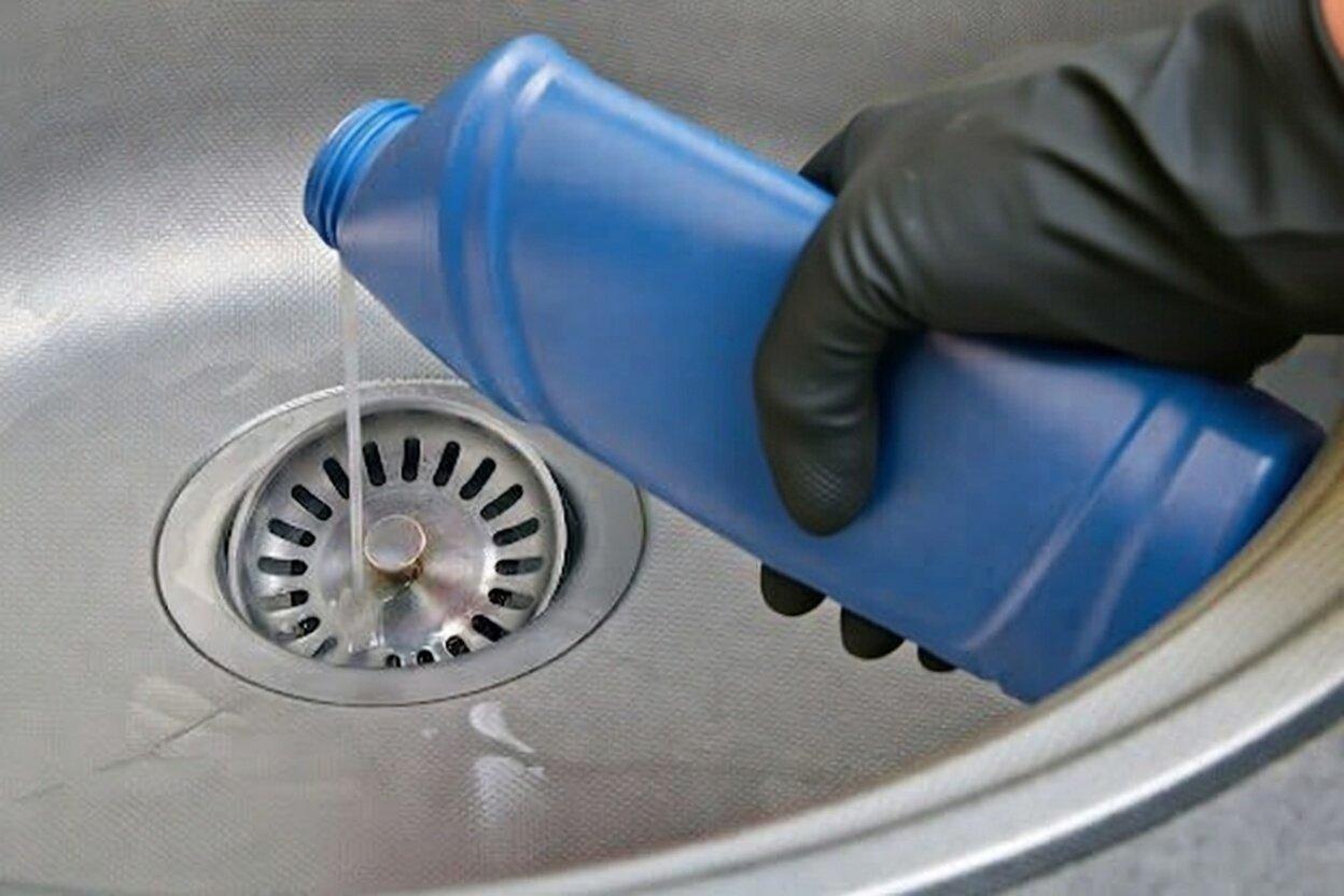 A person wearing a black glove pouring liquid from a blue plastic bottle into a sink drain.