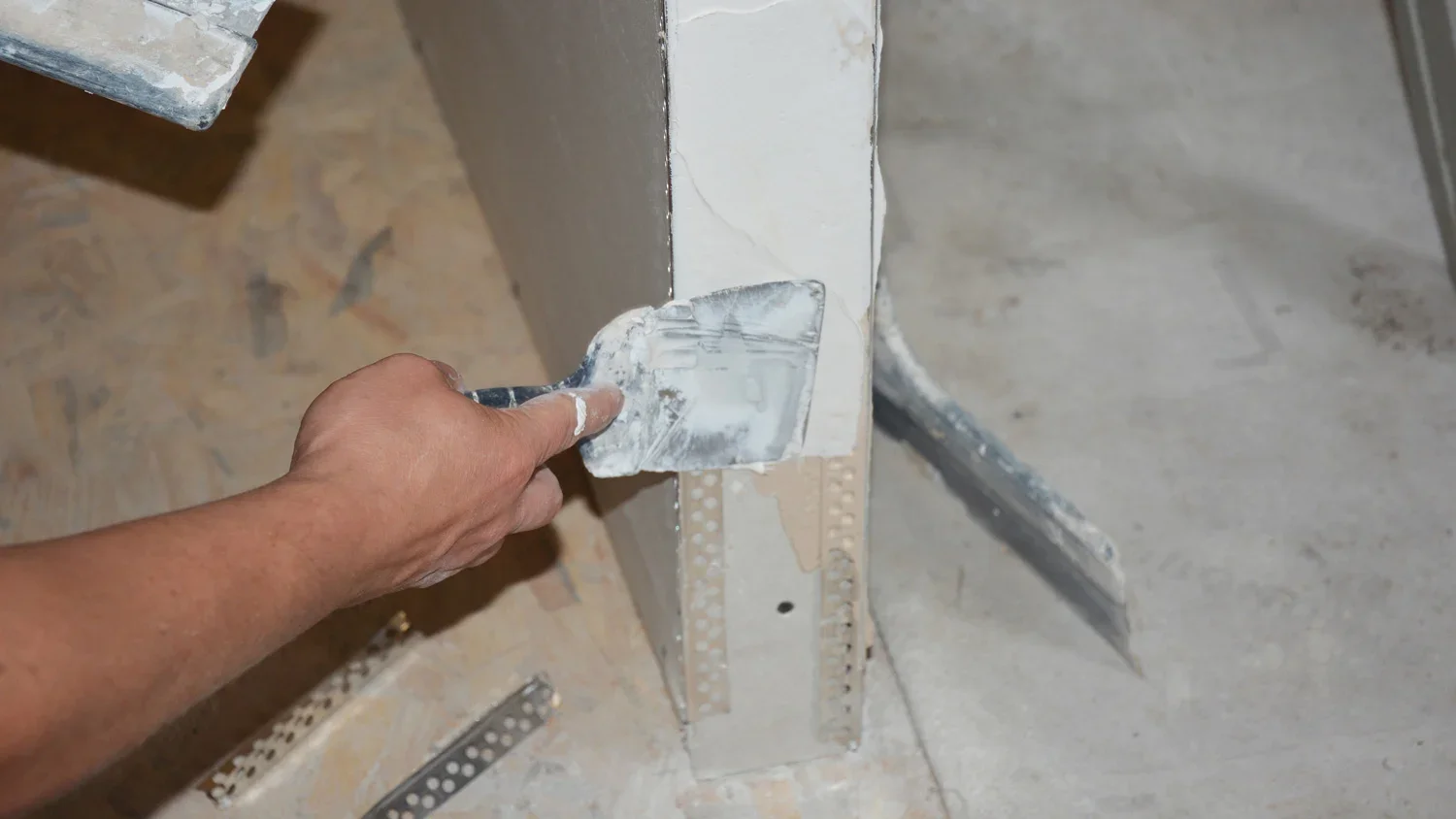 A person is applying joint compound with a putty knife to a vertical corner of drywall during construction or renovation.