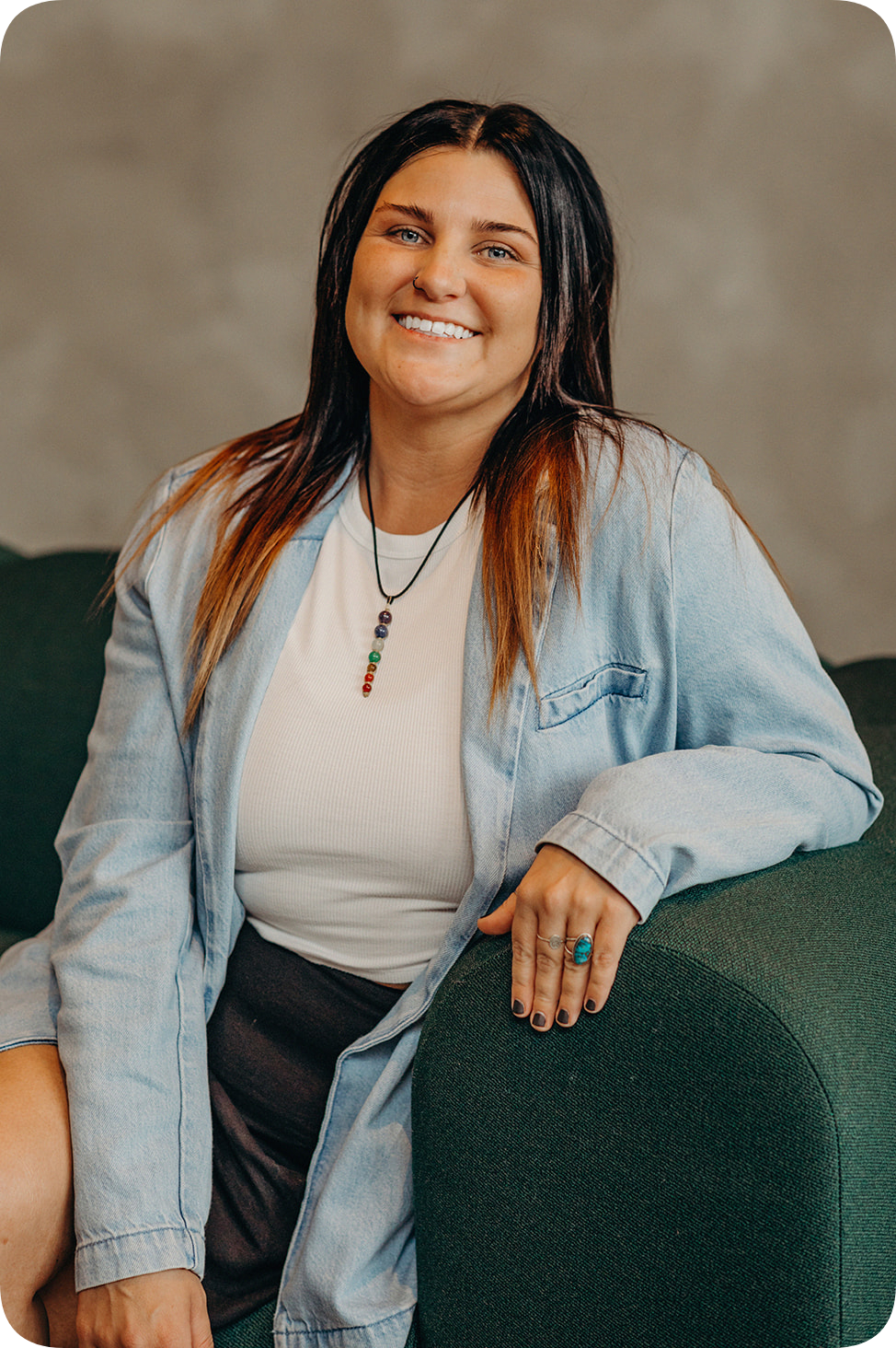 A smiling woman with dark hair, wearing a white shirt, leather skirt, and a light denim jacket, sitting on a dark green couch.
