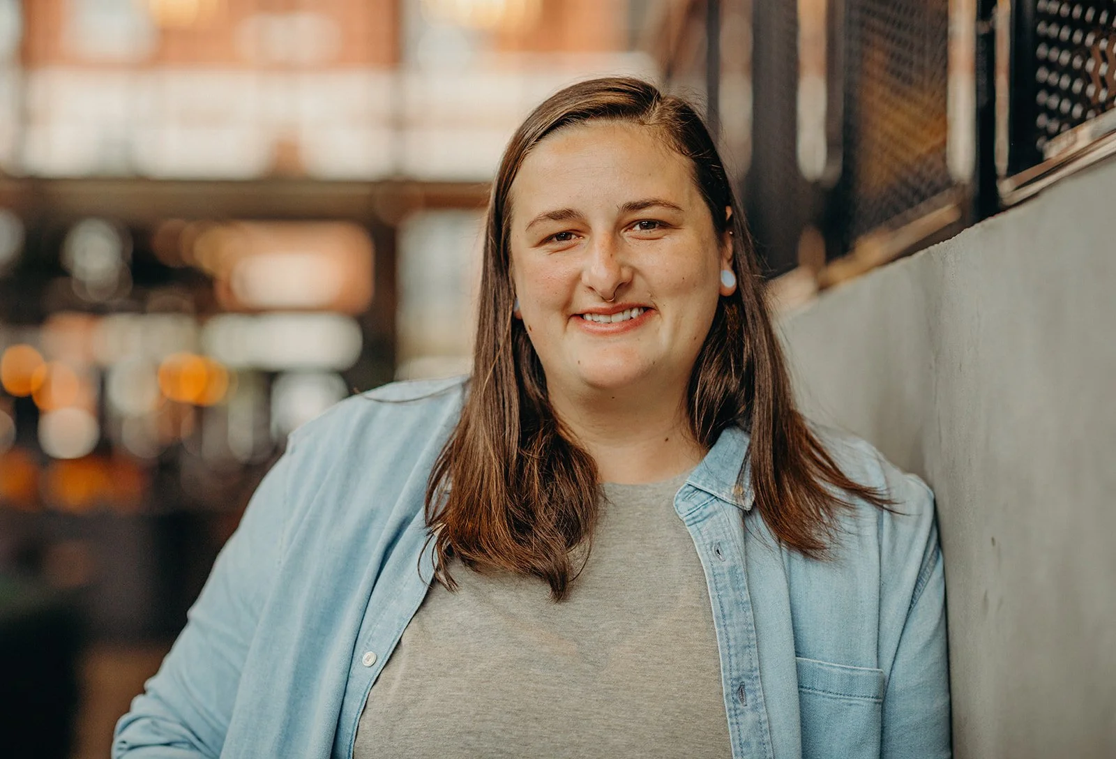 A young woman with long brown hair, wearing a light blue denim shirt and a grey t-shirt, smiling while leaning against a wall in an indoor setting with blurred warm lighting in the background.