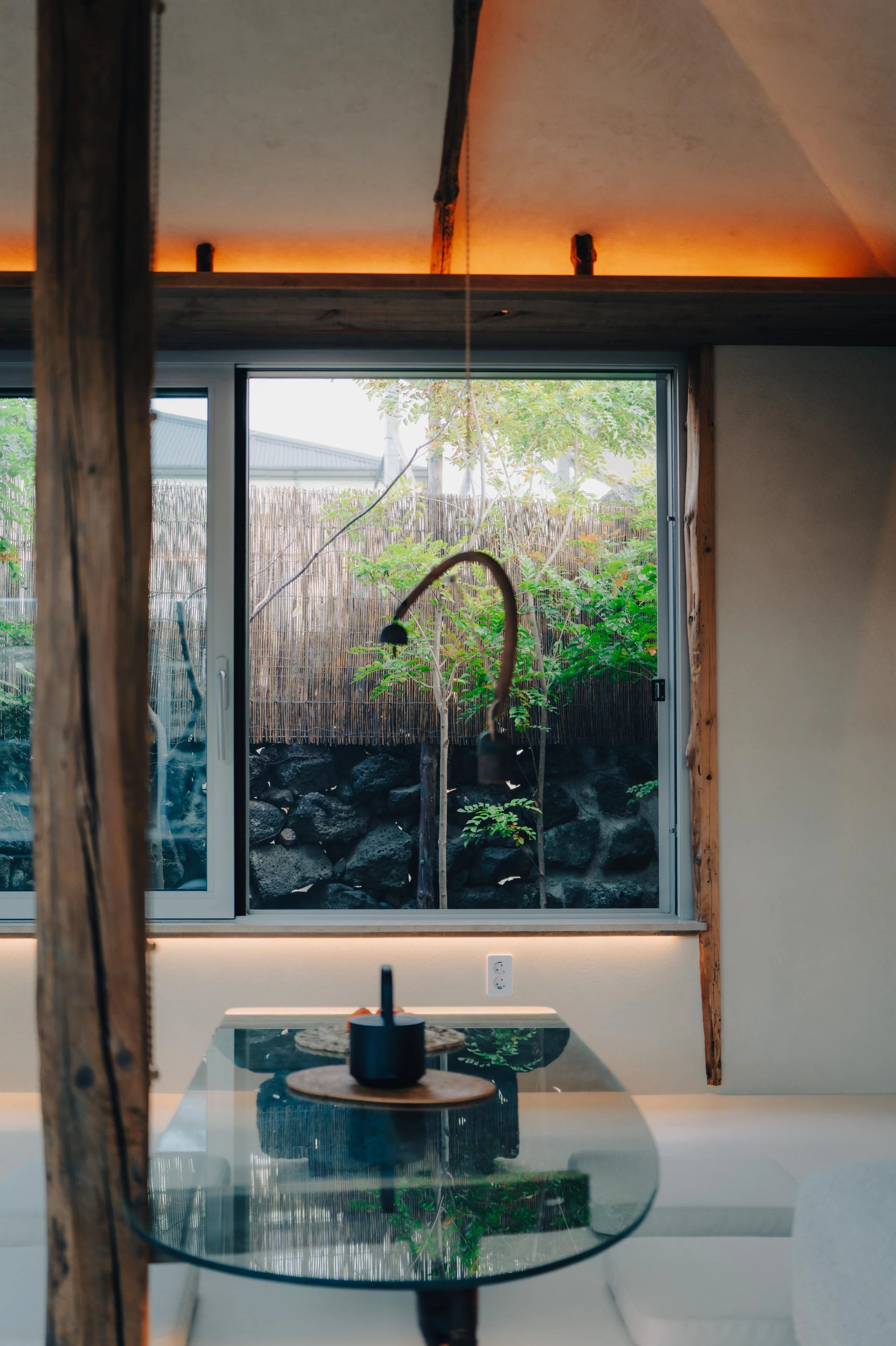 NUUT Aewol 눗애월 Interior view of a modern room with a glass table, a lamp, a large window revealing a small garden with trees and rocks outside, and warm ceiling lighting.